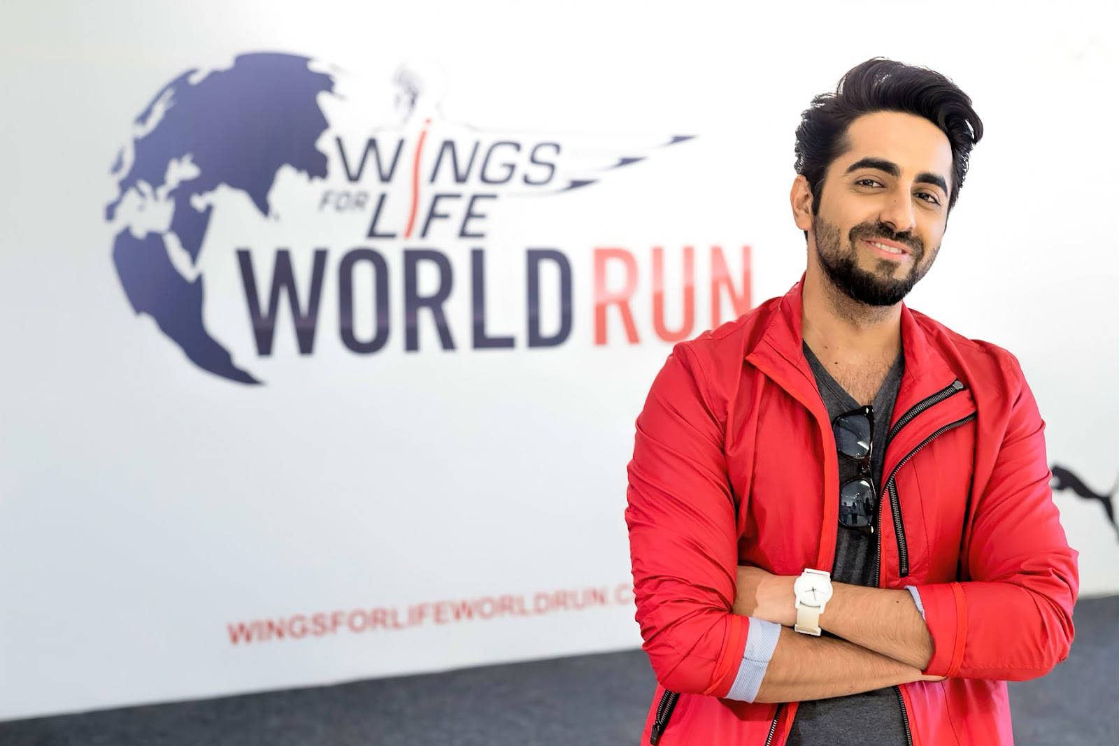ayushmann khurrana 桌布 壁紙 — 一位身穿紅色外套、面帶微笑的男子自信地交叉雙臂，站在「Wings for Life World Run」活動背景前。
