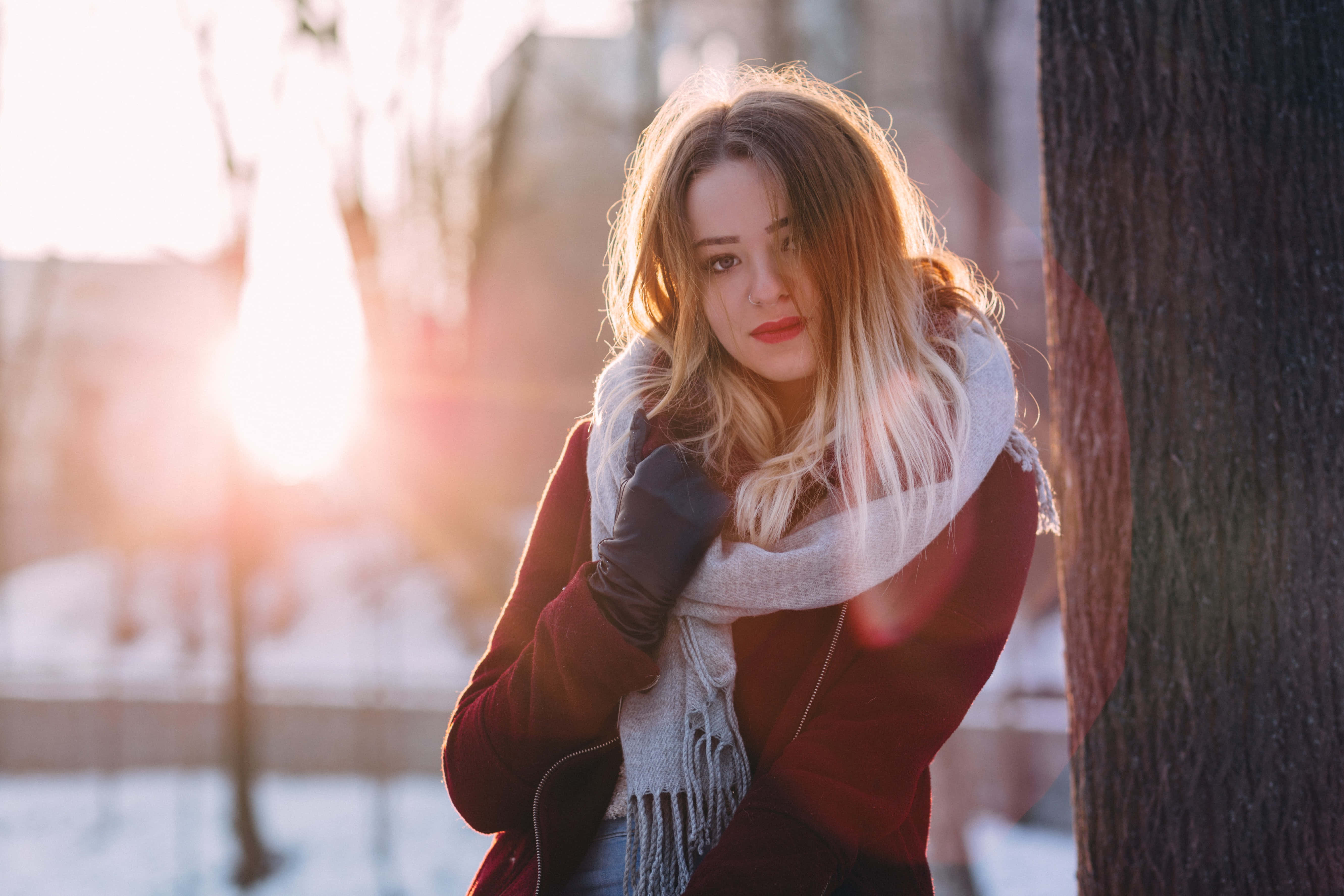 美女 桌布 壁紙 — 一位身穿紅色外套和白色圍巾的女性靠在雪地公園的樹上，身後金色陽光閃耀。