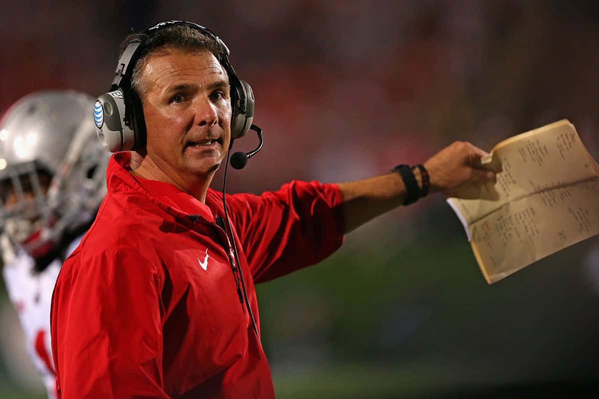 Urban Meyer 桌布 壁紙 — 一位身穿紅色外套和耳機的足球教練在夜間比賽中，在體育場燈光下用手勢指示戰術板。