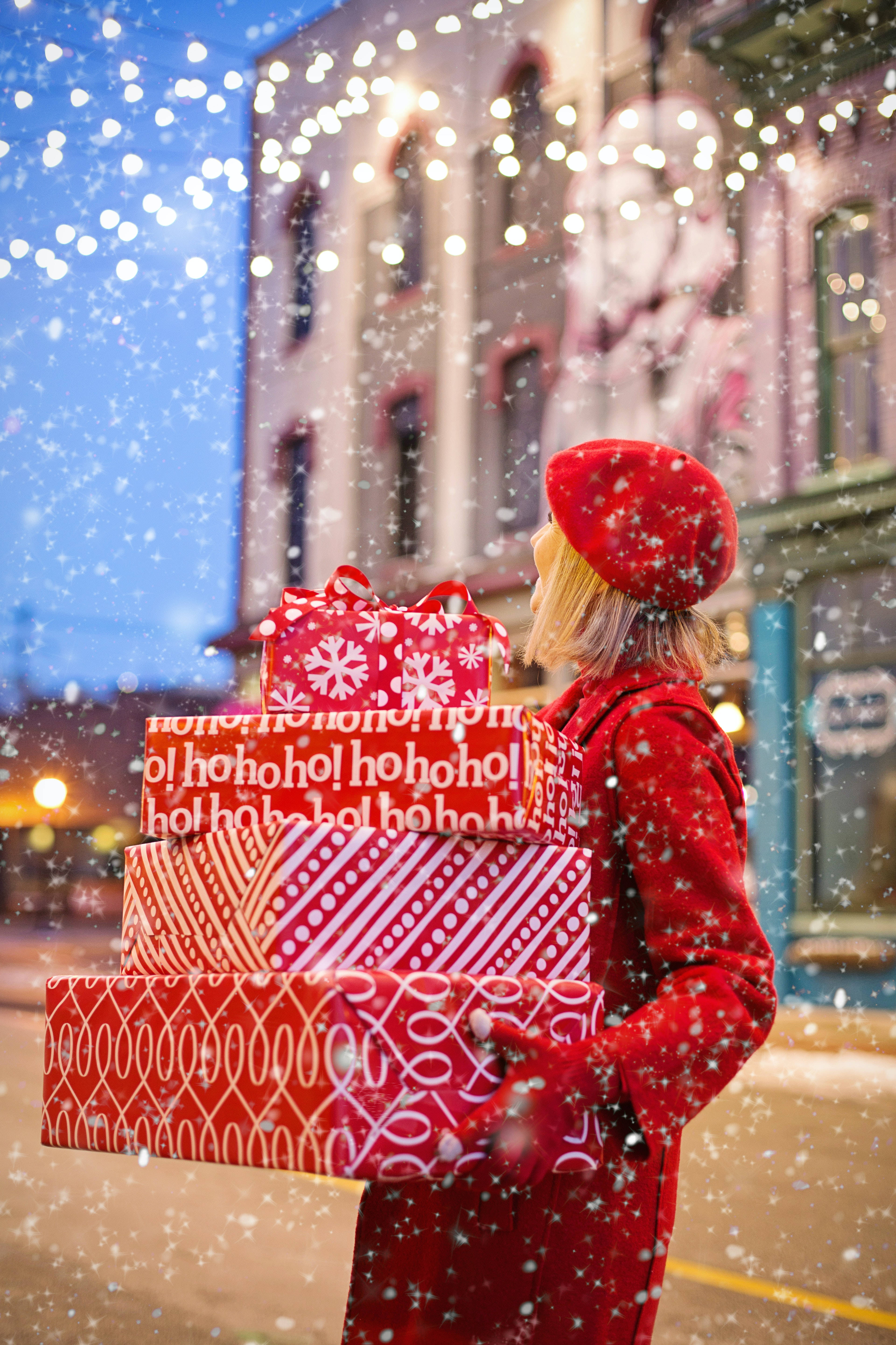 Snowy Christmas 桌布 壁紙 — 一位身穿紅色外套和貝雷帽的女性，在黃昏時分溫暖照明的城市街道上，雪花飄落，她抱著一疊節日紅色禮物盒。