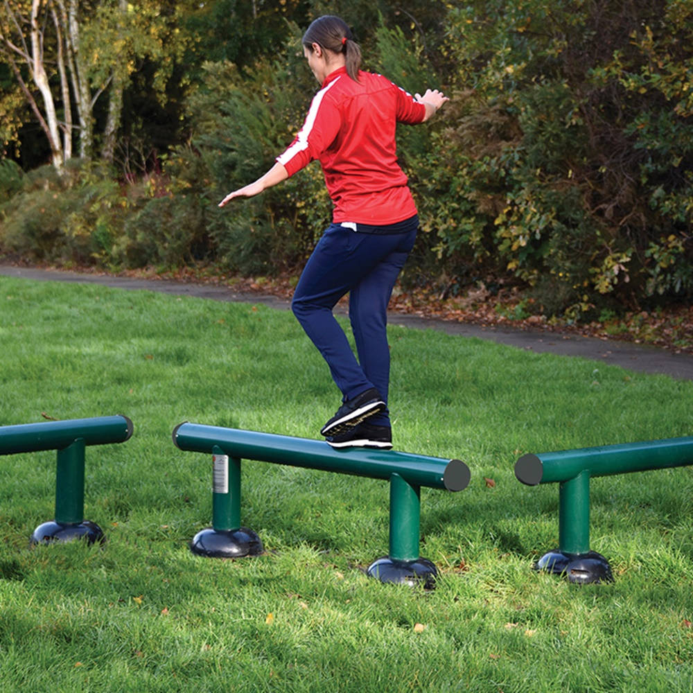 Exercise 桌布 壁紙 — 一位身穿紅色外套的女性在一個被秋樹環繞的草地公園中的綠色戶外橫桿上保持平衡。