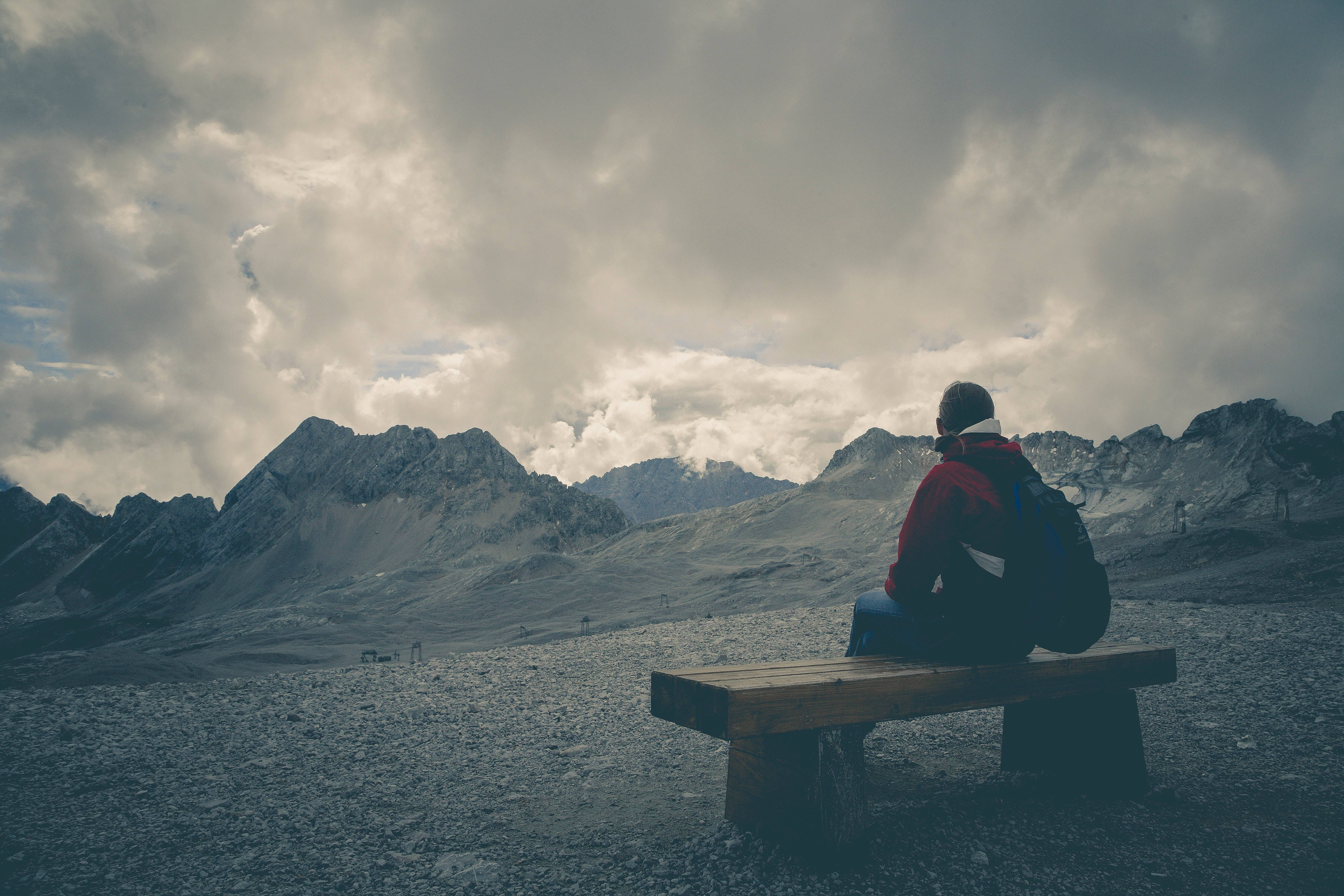 Alone 桌布 壁紙 — 一位身穿紅色外套的孤獨遠足者坐在木長椅上，俯瞰戲劇性多雲天空下崎嶇的雪山。