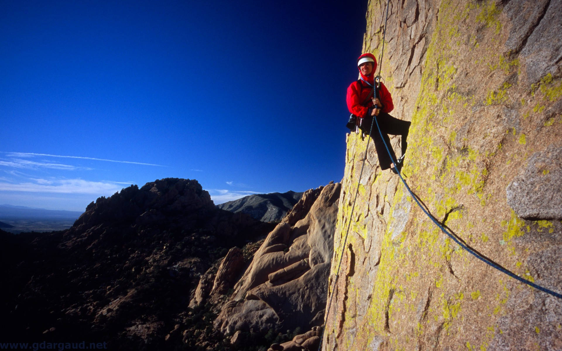 Climbing 桌布 壁紙 — 一位身穿紅色外套的攀岩者，在廣闊的藍天和遠山背景下，攀登著一處有黃色地衣的陽光照射的岩壁。