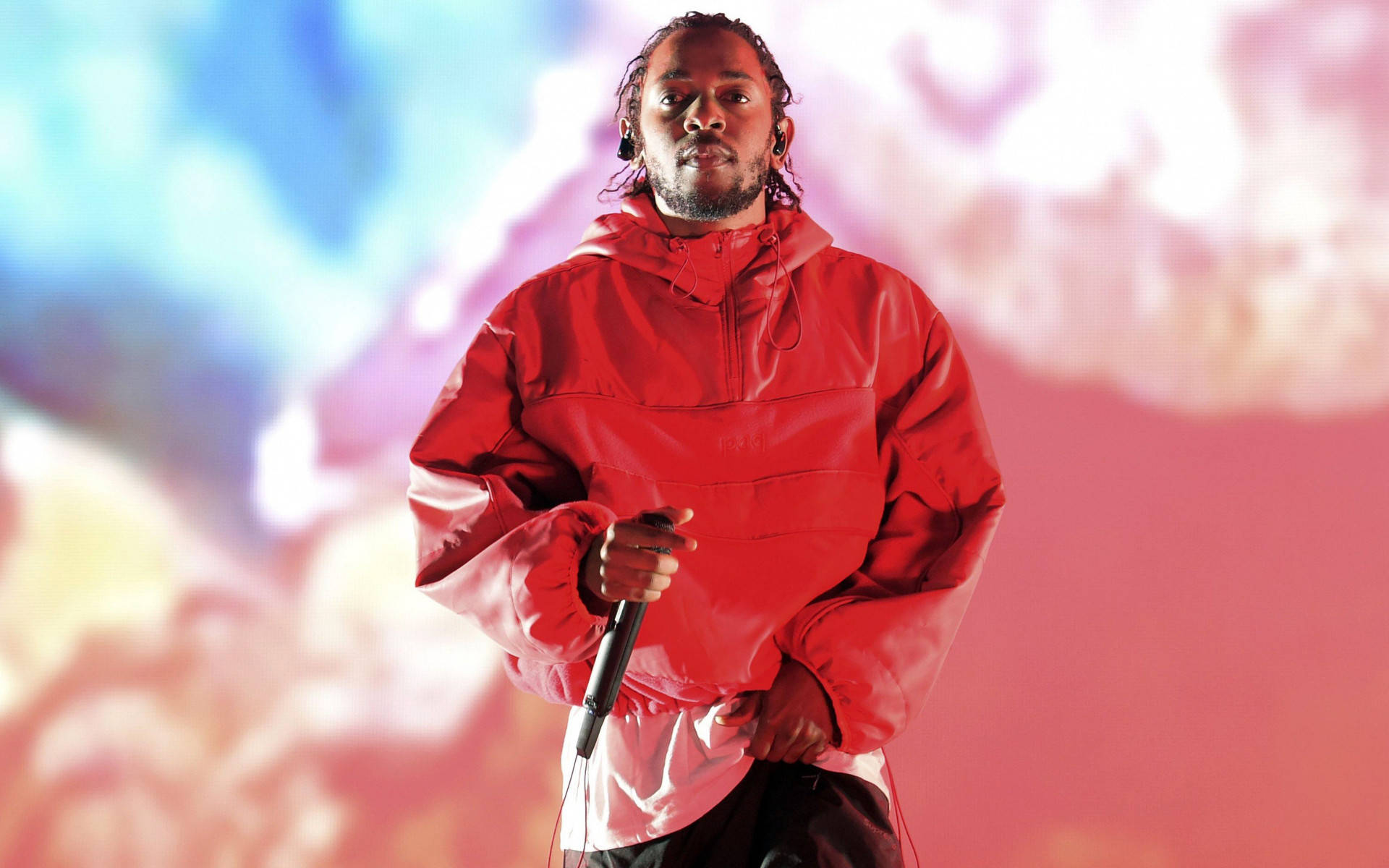 Kendrick 桌布 壁紙 — 一位身穿紅色外套的男性表演者，在粉紅色、藍色和白色雲朵的鮮豔抽象背景前握著麥克風。