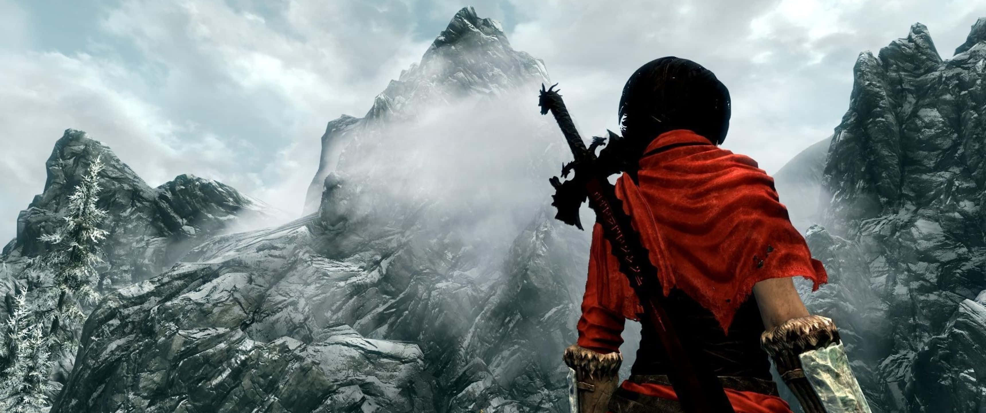 3440x1440p The Elder Scrolls V Skyrim 桌布 壁紙 — 一位身穿紅色斗篷的兜帽戰士站在霧氣瀰漫的雪山山脈前，手持一把劍。