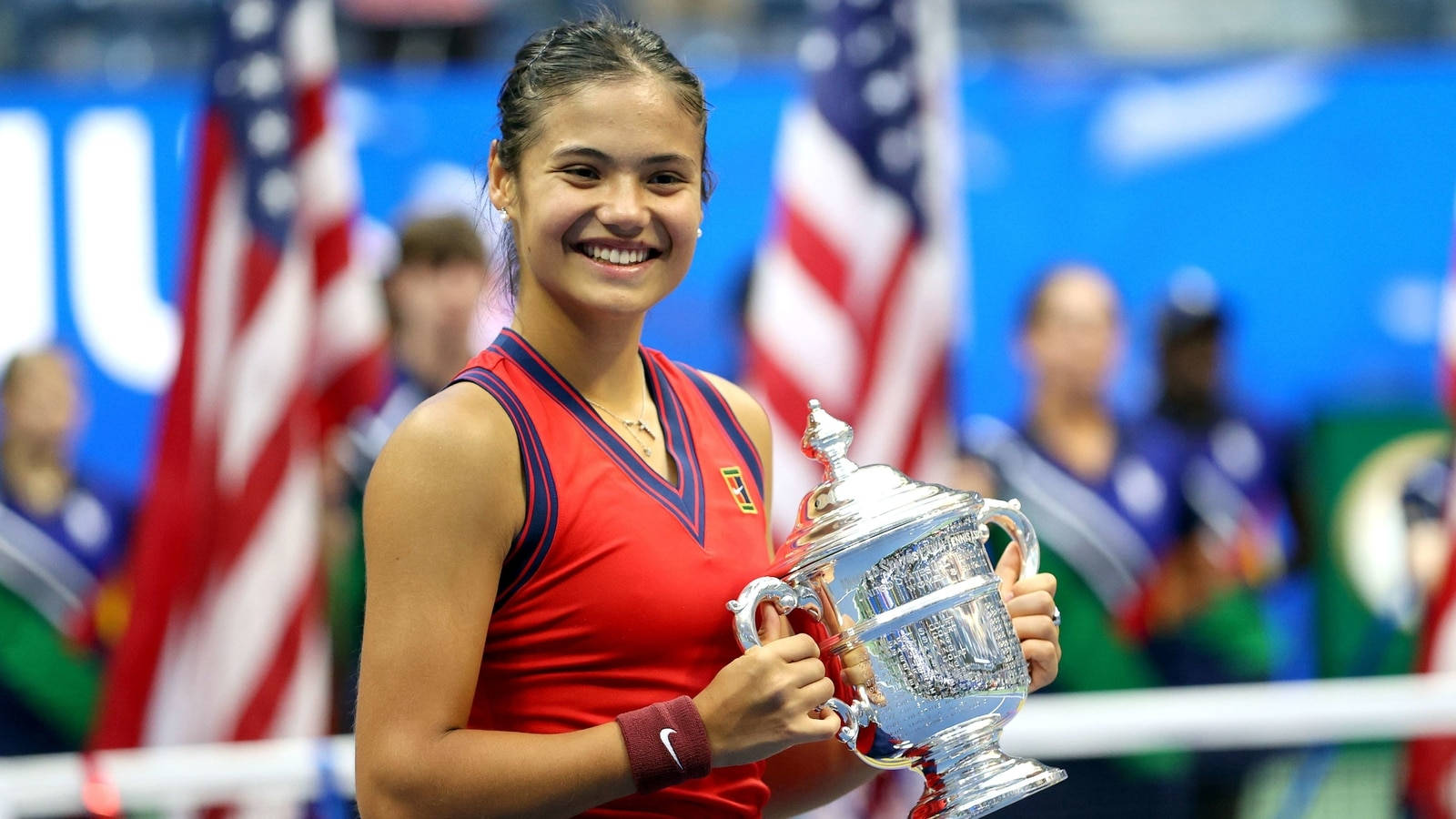 Emma 桌布 壁紙 — 一位身穿紅色服裝、面帶微笑的女性網球選手，身後有美國國旗，自豪地高舉一個大型銀色獎盃。