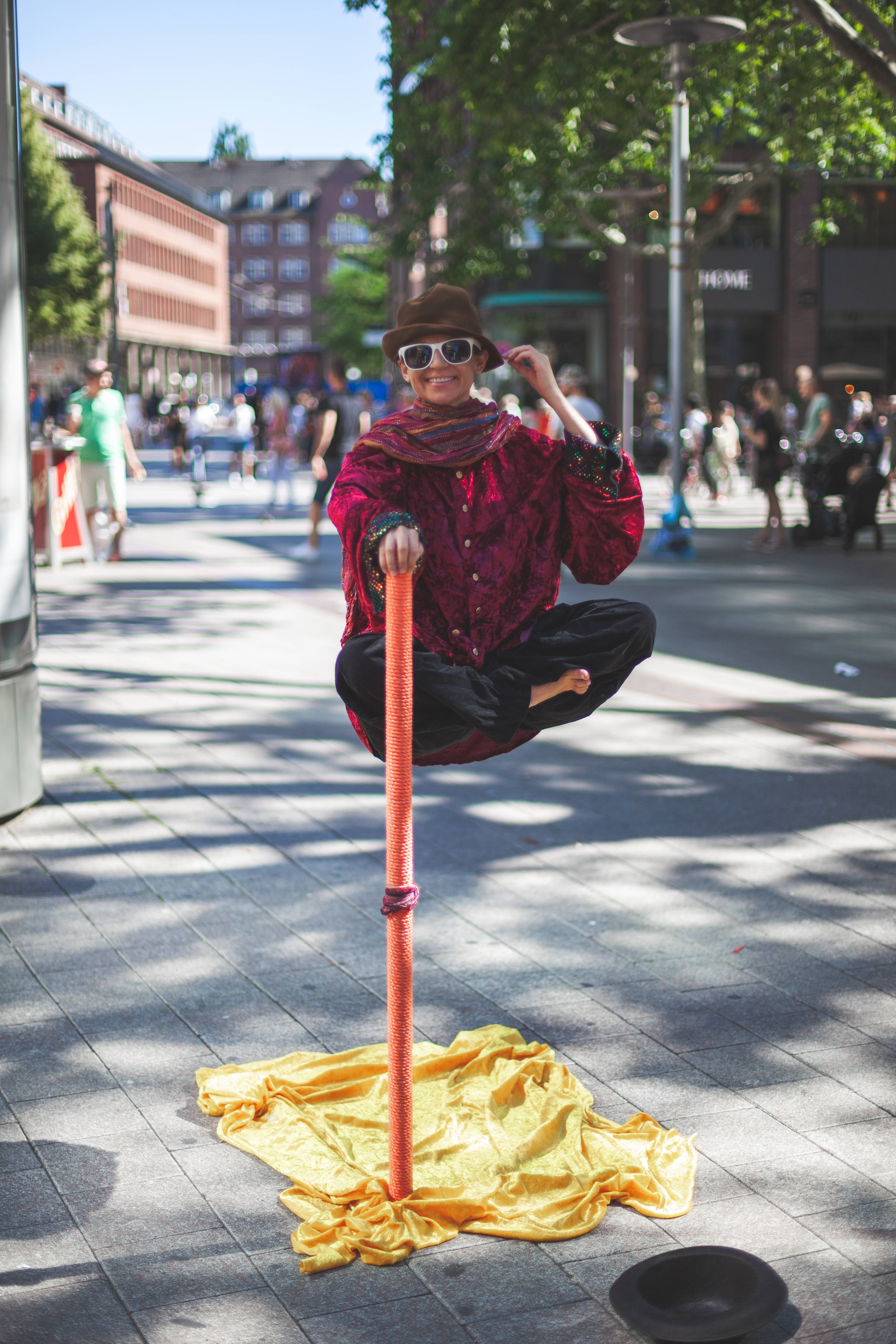 Illusion 桌布 壁紙 — 一位身穿紅色服裝和帽子的街頭表演者在陽光明媚的城市廣場上優雅地平衡在桿子上，同時對著鏡頭微笑。