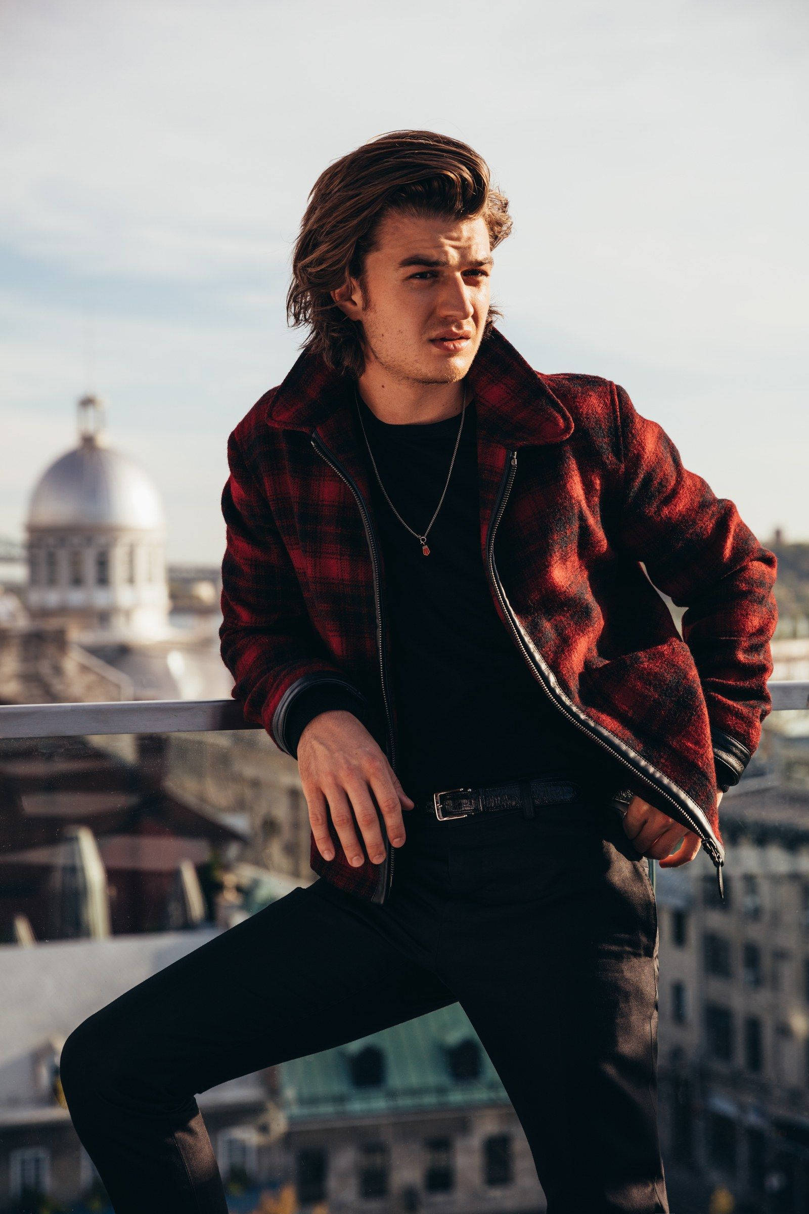 Joe Keery 桌布 壁紙 — 一位身穿紅色格子外套的年輕男子自信地靠在屋頂欄杆上，身後是城市天際線和圓頂建築，天空晴朗。