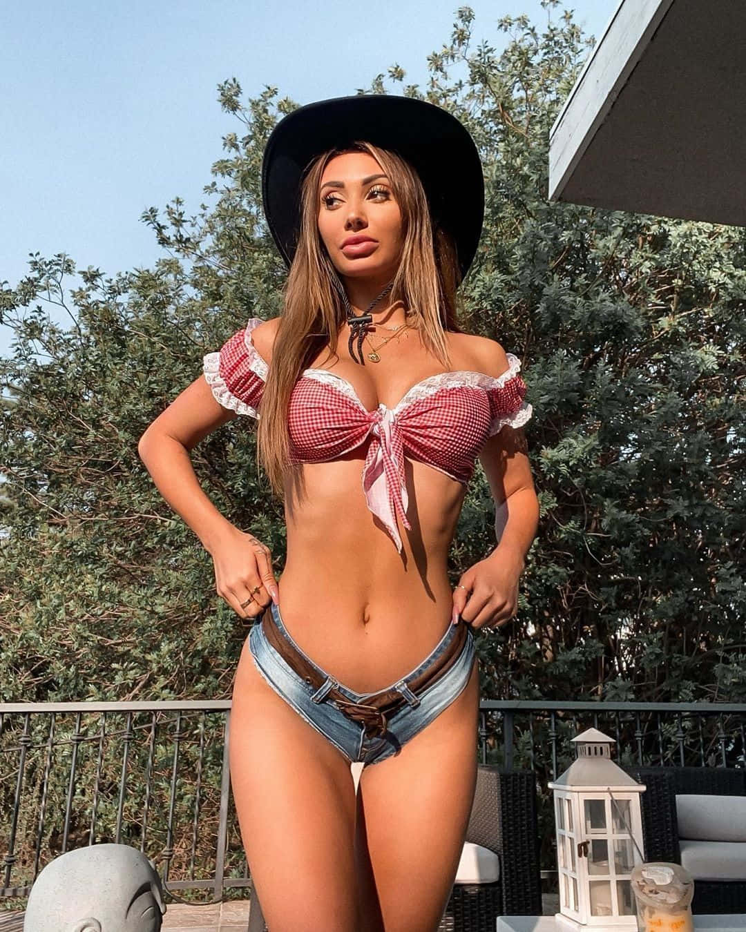 sexy bikini 桌布 壁紙 — 一位身穿紅色格紋上衣和牛仔短褲的女性，在明亮的日光下，於被綠色植物環繞的陽台上自信地擺姿勢。