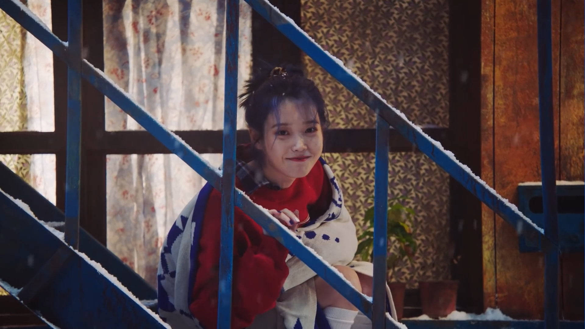 Cute Girl 桌布 壁紙 — 一位身穿紅色毛衣和圖案毯子的年輕女性，坐在室內灑滿雪的藍色樓梯上，溫柔地微笑。