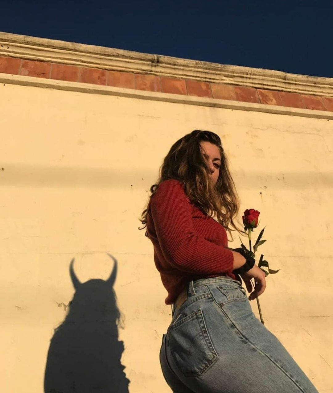 Rose Flower 桌布 壁紙 — 一位身穿紅色毛衣和牛仔褲的女性，在陽光照射的牆邊舉著一朵玫瑰，她的影子投射出惡魔的角。