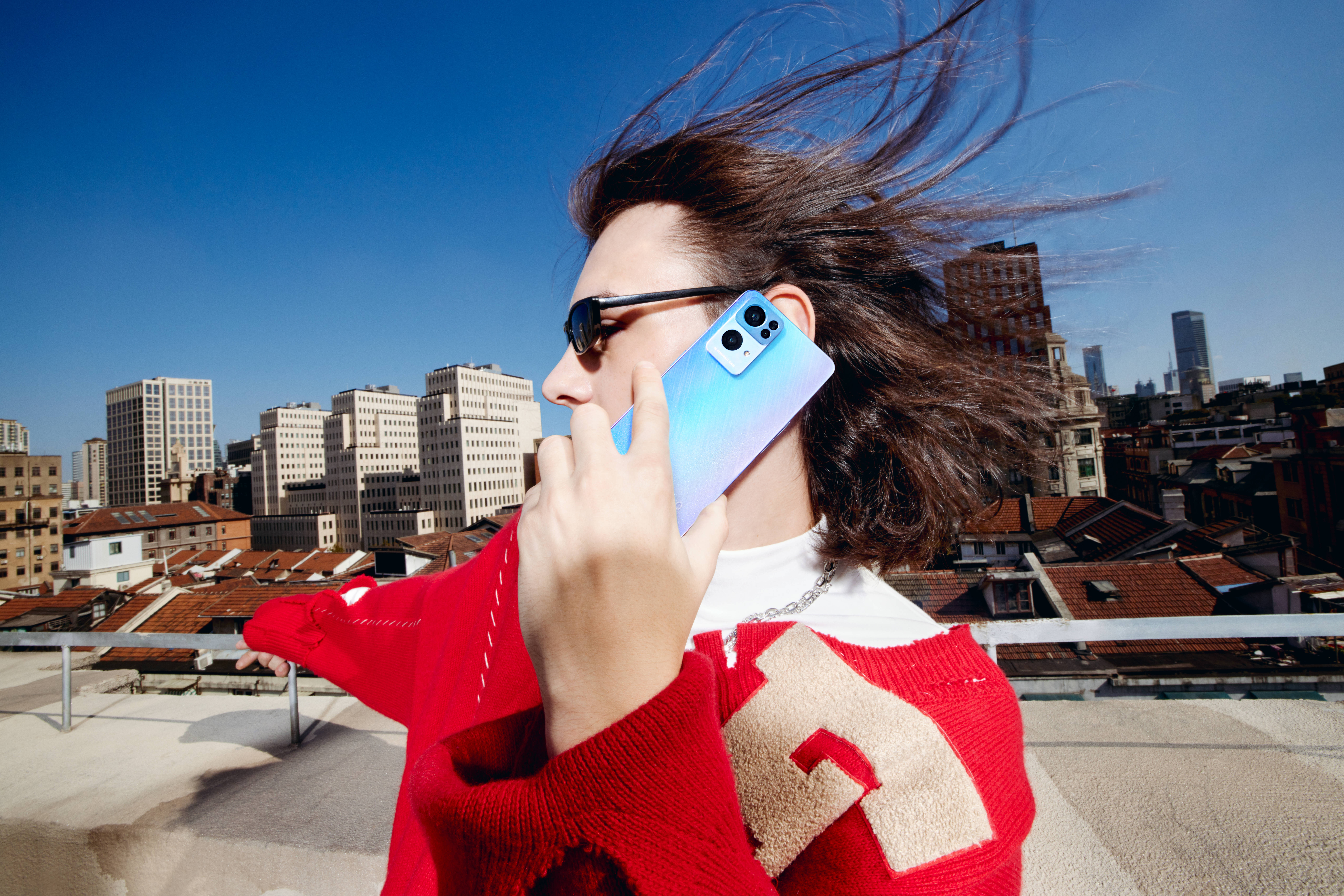 oppo 桌布 壁紙 — 一位身穿紅色毛衣的女性在都市屋頂上使用藍色電話通話，風吹動她的頭髮，背景是清澈的藍天。