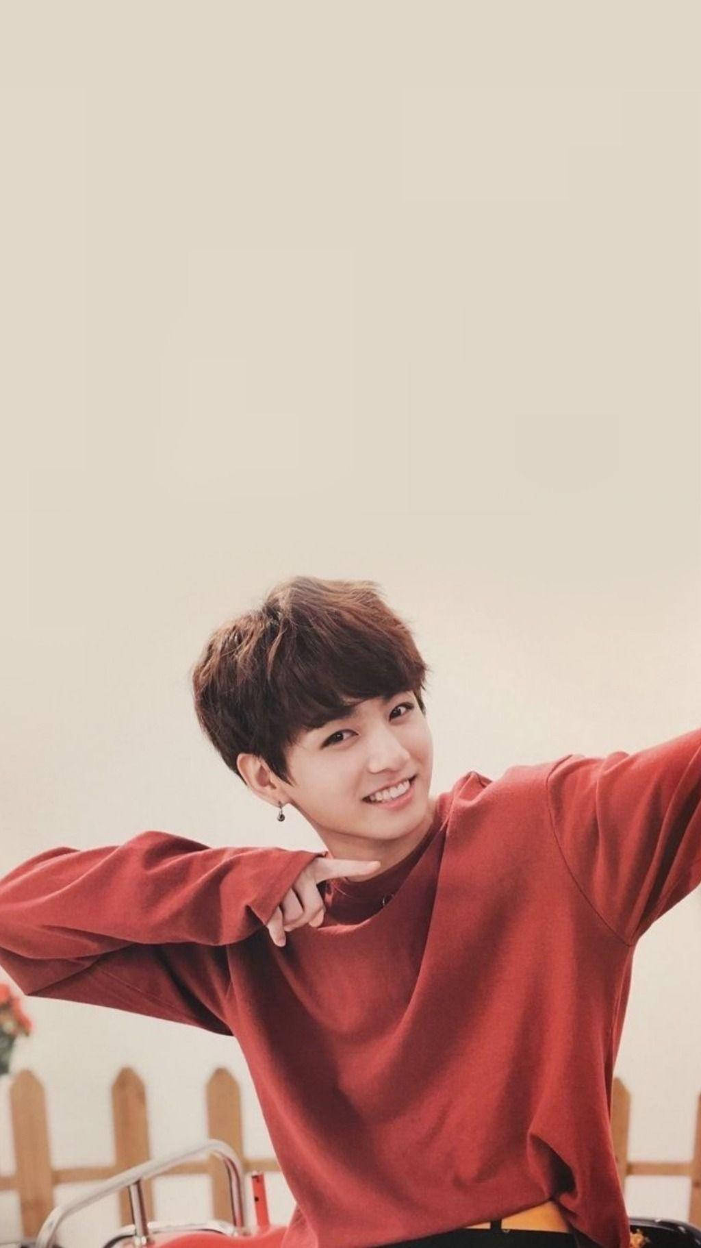 Jungkook Aesthetic 桌布 壁紙 — 一位身穿紅色毛衣的年輕男子，在柔和的米色背景前張開雙臂，露出燦爛的笑容。