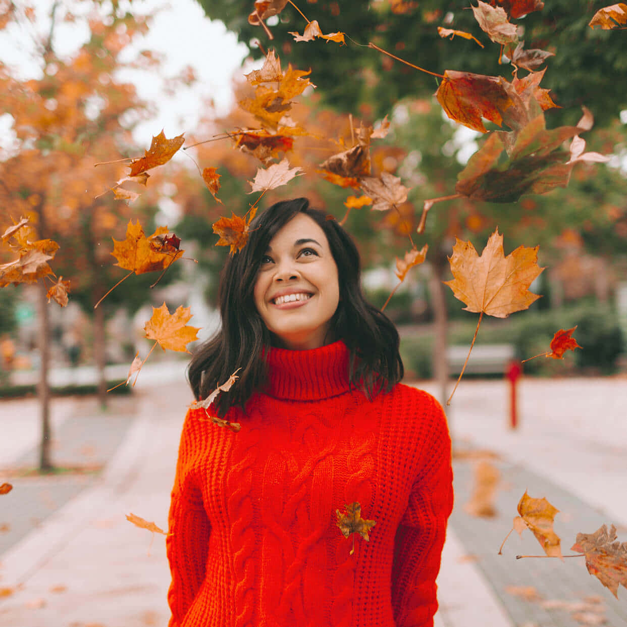 Autumn Fall 桌布 壁紙 — 一位身穿紅色毛衣的快樂女性在充滿活力的秋季公園場景中微笑，秋葉在她周圍旋轉。