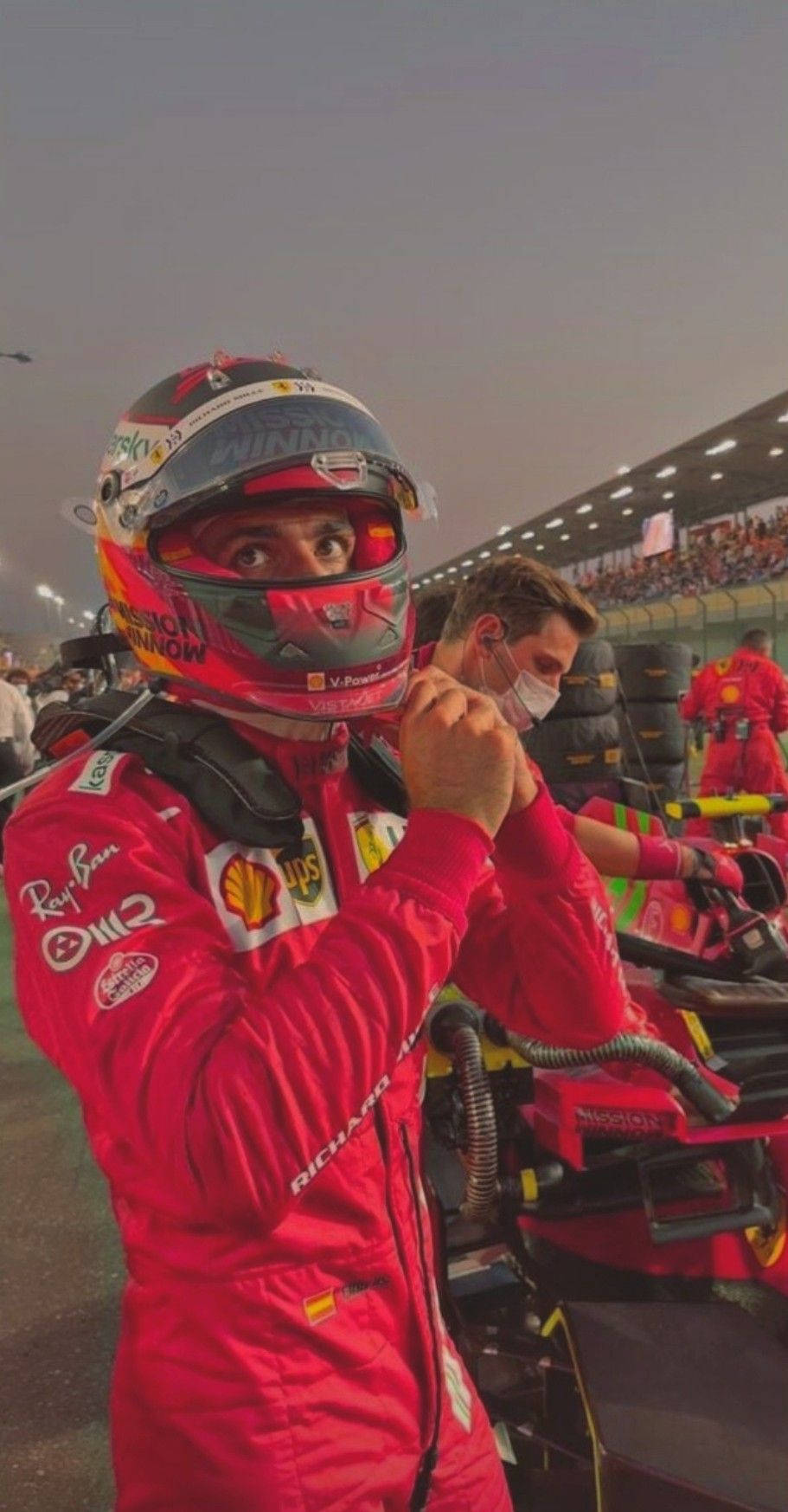 Racing 桌布 壁紙 — 一位身穿紅色法拉利賽車服的F1車手在黃昏的賽道上調整頭盔，背景有維修站工作人員和觀眾。