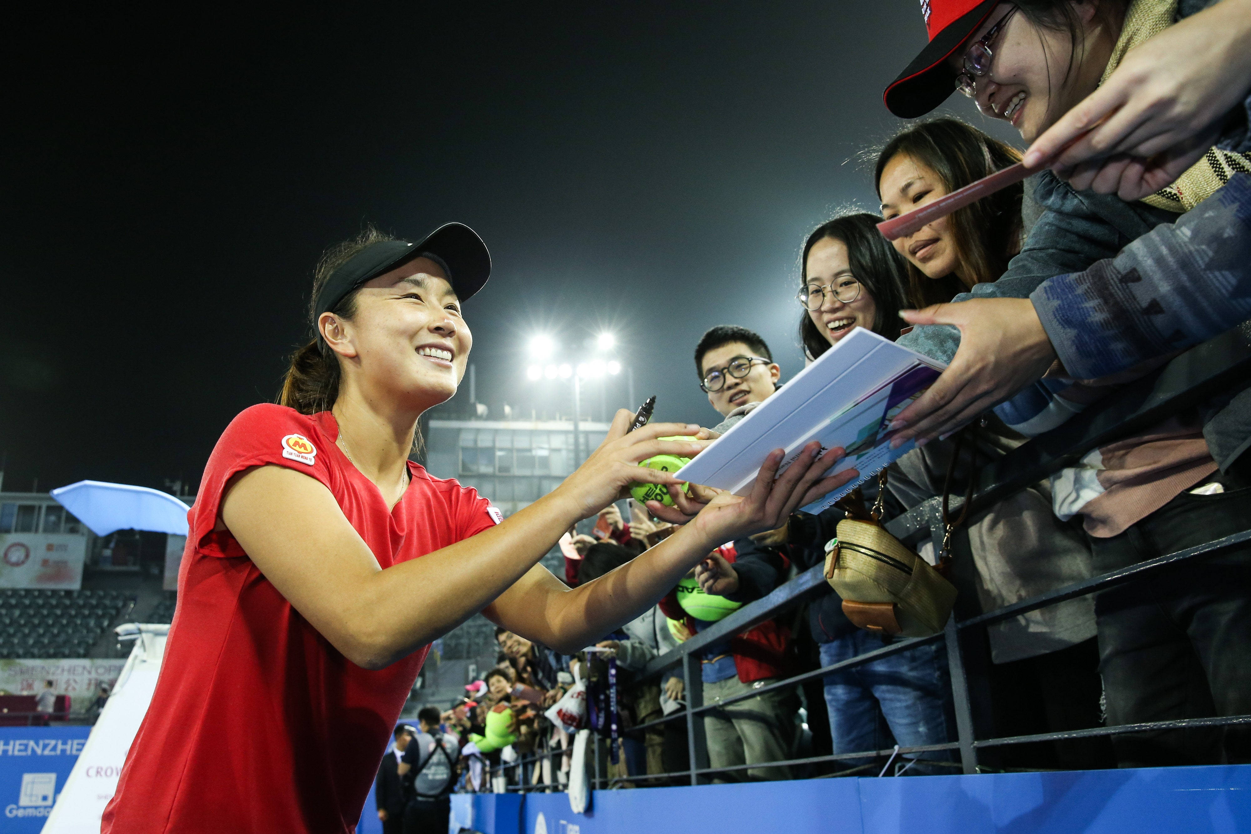 網球 桌布 壁紙 — 一位身穿紅色球衣、面帶微笑的女網球選手在夜間明亮的體育場燈光下為球迷簽名。