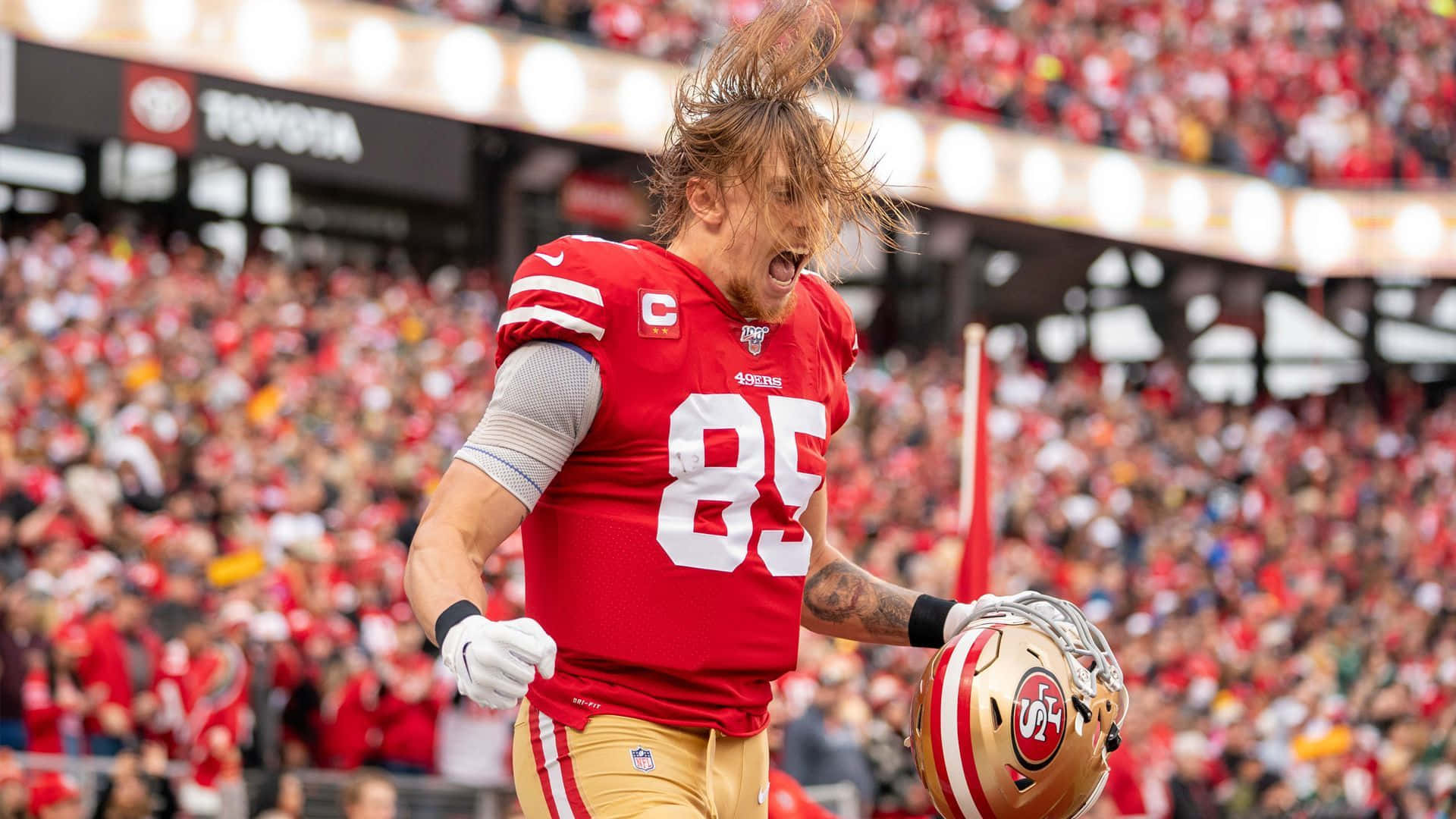 George Kittle 桌布 壁紙 — 一位身穿紅色球衣85號的舊金山49人美式足球員，在擠滿觀眾的球場上，手持頭盔，頭髮凌亂地慶祝。