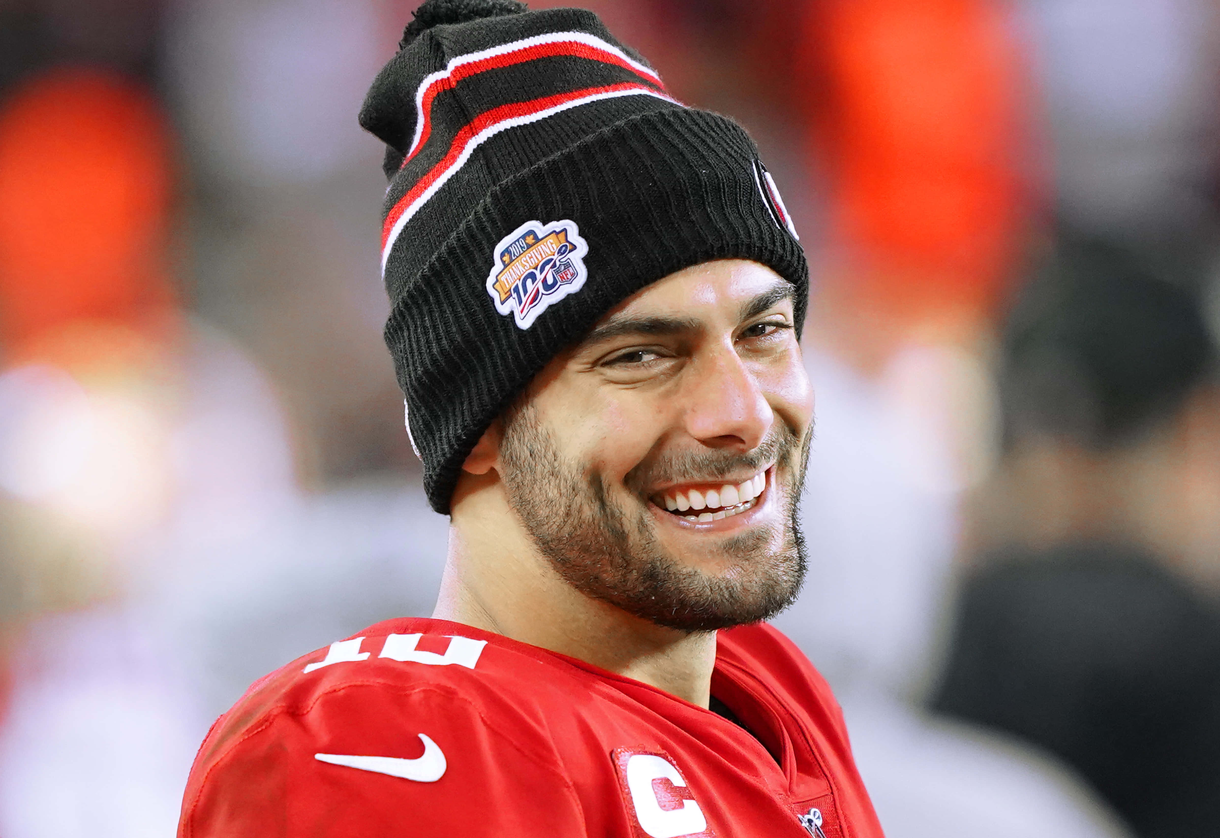 Jimmy Garoppolo 桌布 壁紙 — 一位身穿紅色球衣和帶有冠軍補丁的黑色毛帽的足球運動員微笑著看向鏡頭外，身後是明亮的體育場燈光。