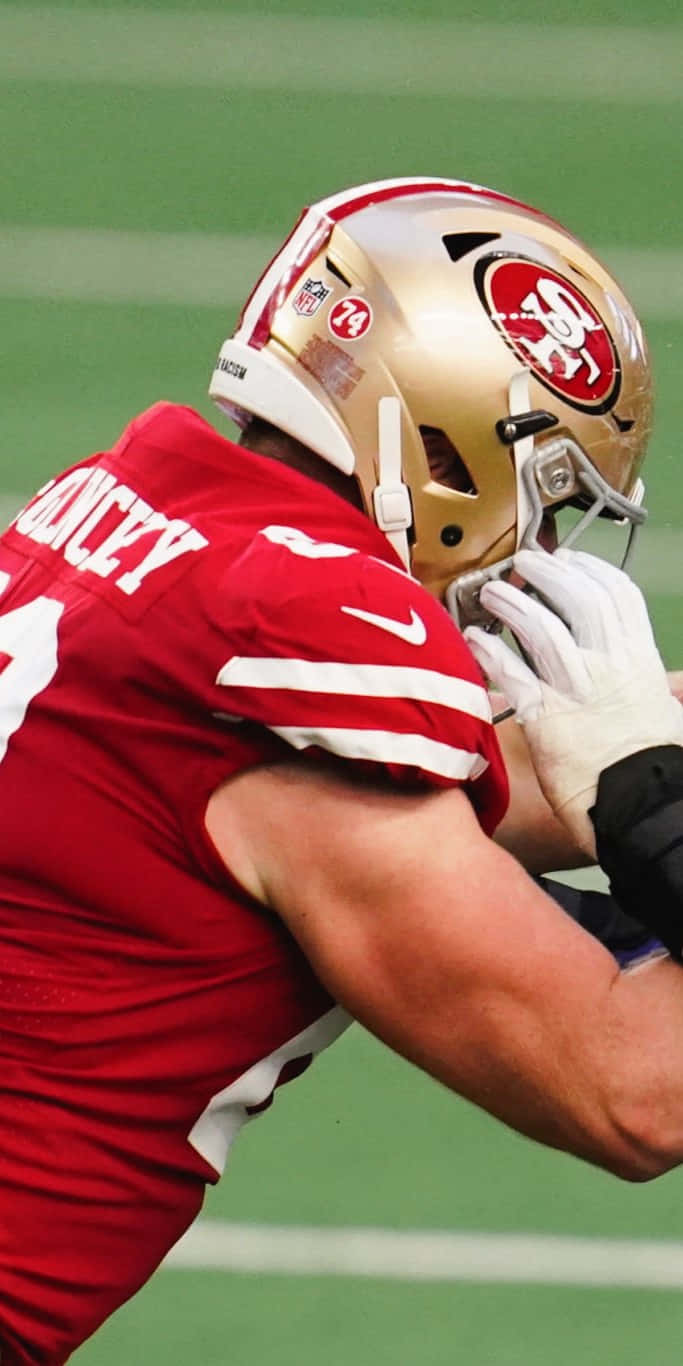 Mike Mcglinchey 桌布 壁紙 — 一位身穿紅色球衣和金色頭盔的舊金山49人美式足球員在綠色球場上調整頭盔。