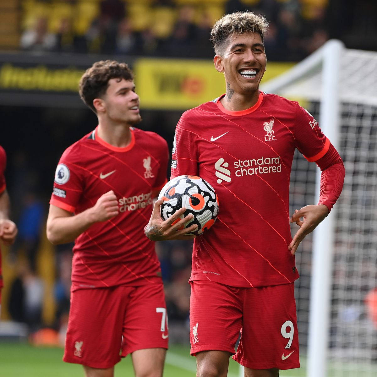 Liverpool Fc 桌布 壁紙 — 一位身穿紅色球衣的利物浦足球員，在綠色球場上拿著比賽用球，身後有球門網，開懷大笑。
