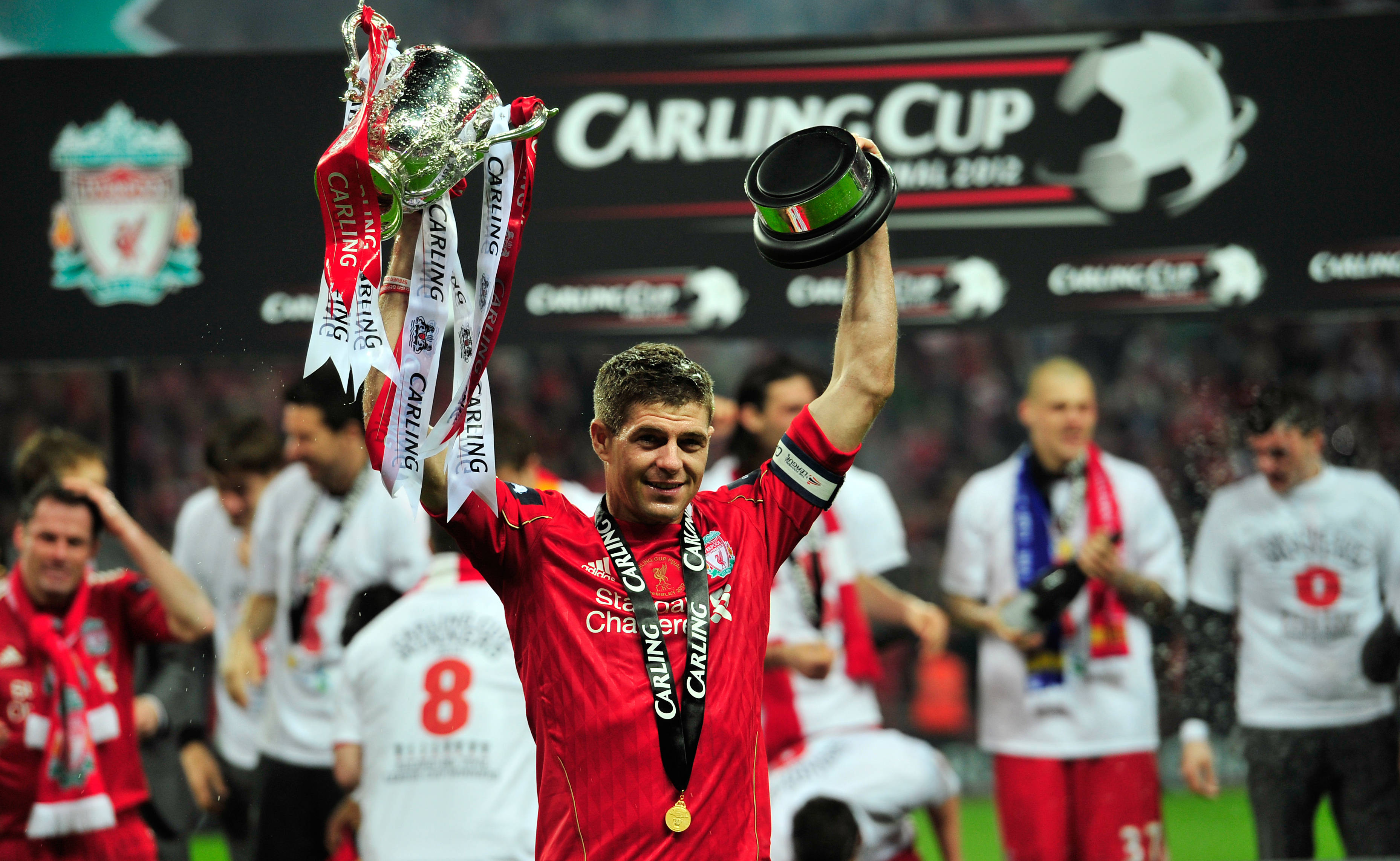 Steven Gerrard 桌布 壁紙 — 一位身穿紅色球衣的利物浦足球員在球場上興奮地高舉閃亮的卡靈盃獎盃和帽子。