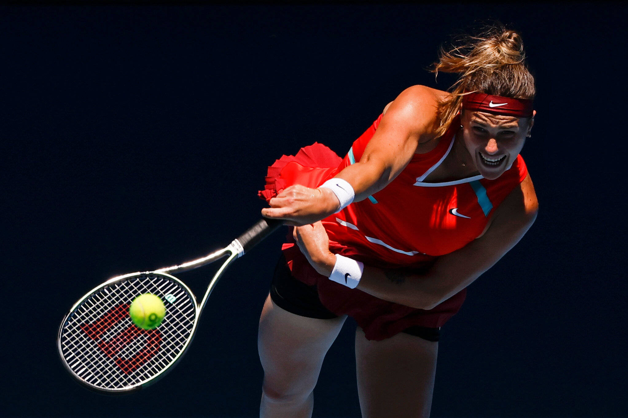 Aryna Sabalenka 桌布 壁紙 — 一位身穿紅色球衣的女網球選手在深色背景下揮拍擊打黃色網球。