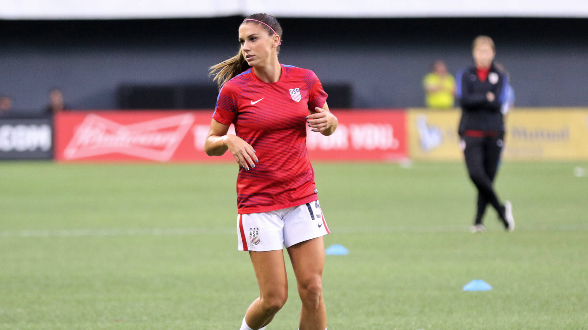 Alex Morgan 桌布 壁紙 — 一位身穿紅色球衣的女足球員在綠色球場上訓練，場上有藍色錐體和模糊的體育場背景。