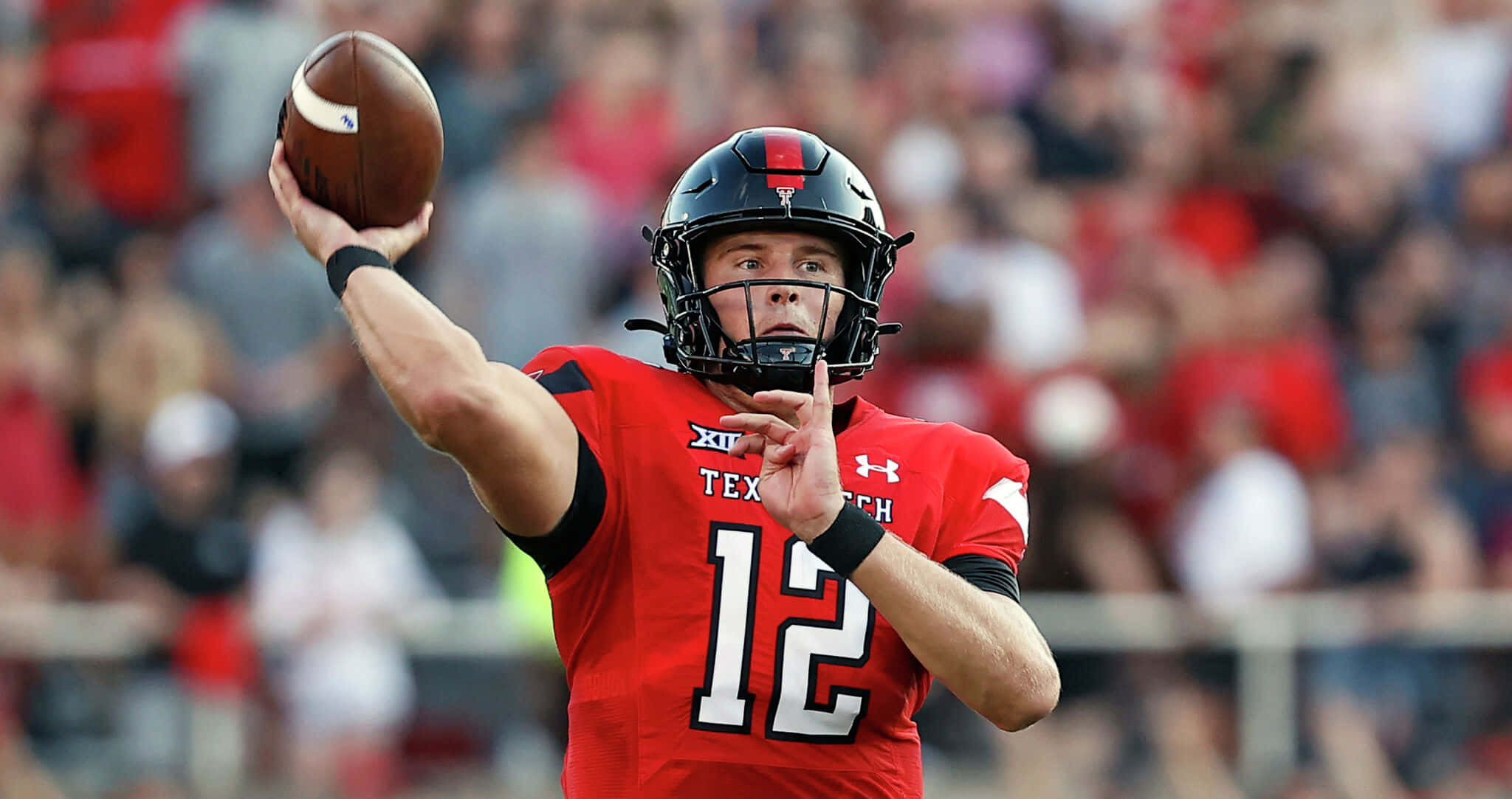 Texas Tech 桌布 壁紙 — 一位身穿紅色球衣的德州理工大學美式足球四分衛在日間比賽中擲出足球，背景是模糊的人群。