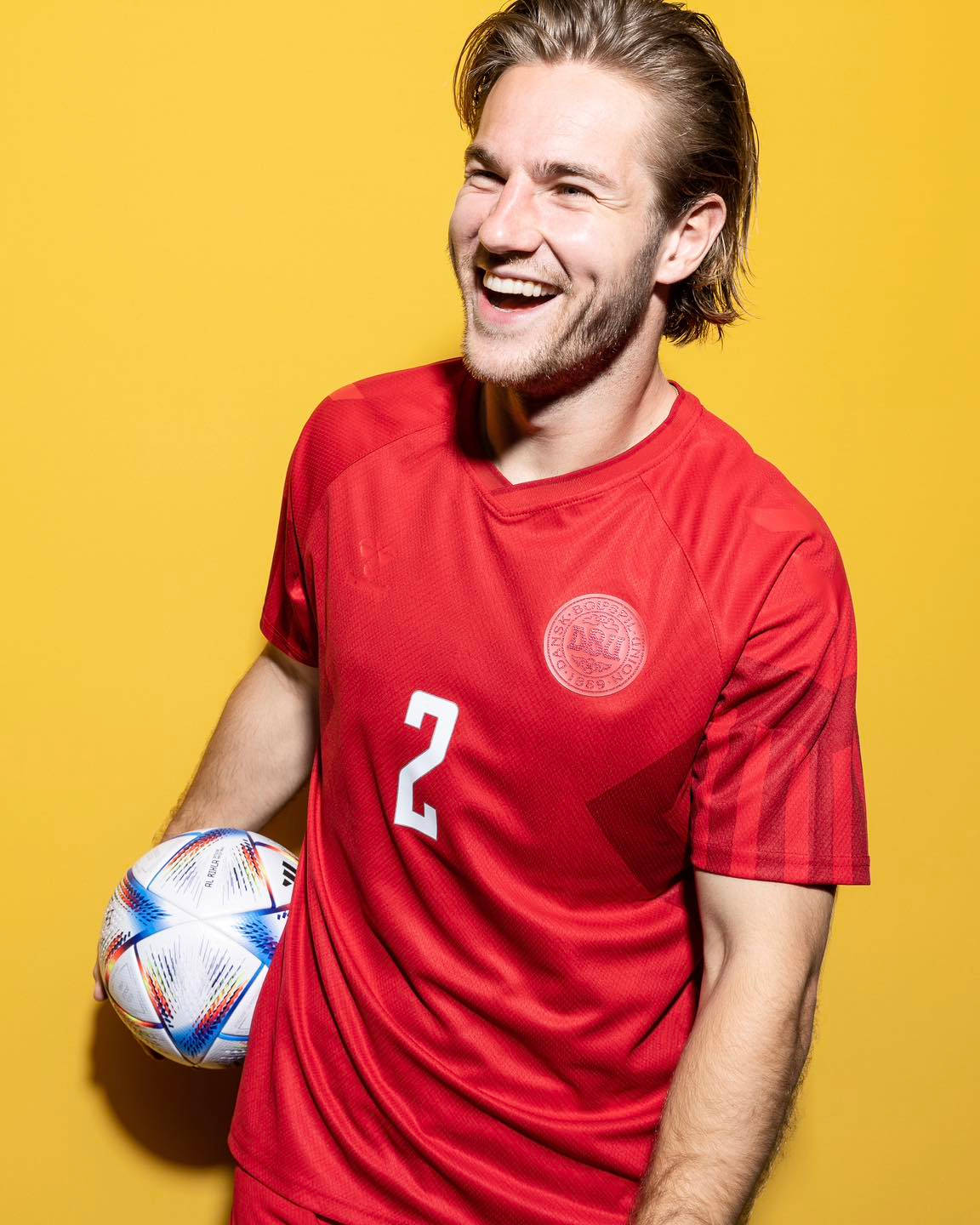 Football Players 桌布 壁紙 — 一位身穿紅色球衣的快樂男足球員，在鮮黃色工作室背景前，笑著拿著一個色彩繽紛的球。