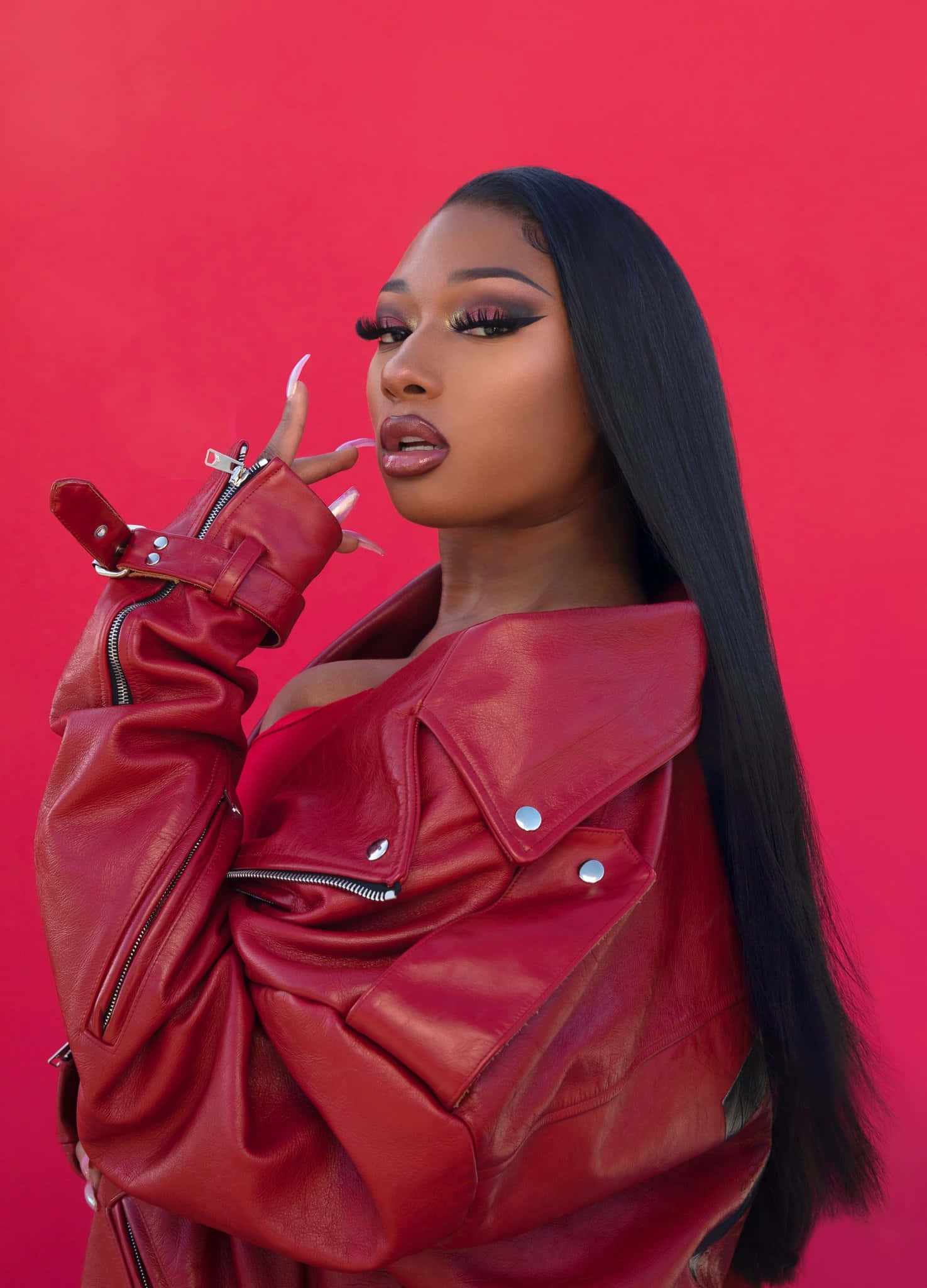 Megan Thee Stallion 桌布 壁紙 — 一位身穿紅色皮夾克的女性，在純紅色背景前自信地擺姿勢，帶有戲劇性的化妝和長長的黑髮。