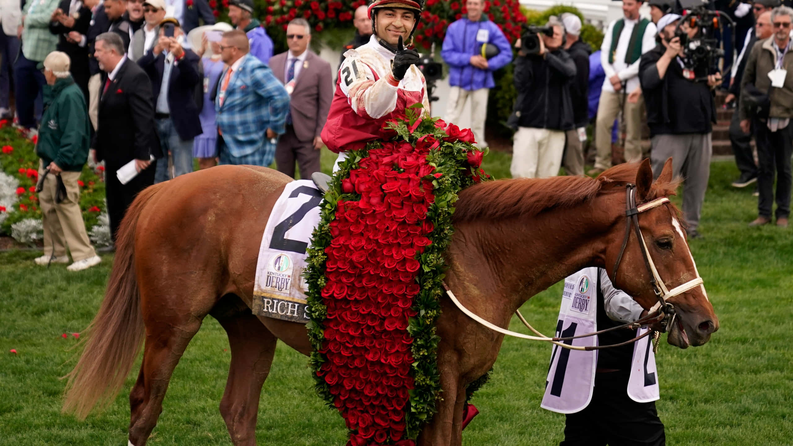 Kentucky Derby 桌布 壁紙 — 一位身穿紅色絲綢的騎師微笑著騎著一匹披著華麗紅玫瑰花環的棕色賽馬，在綠色草地上。