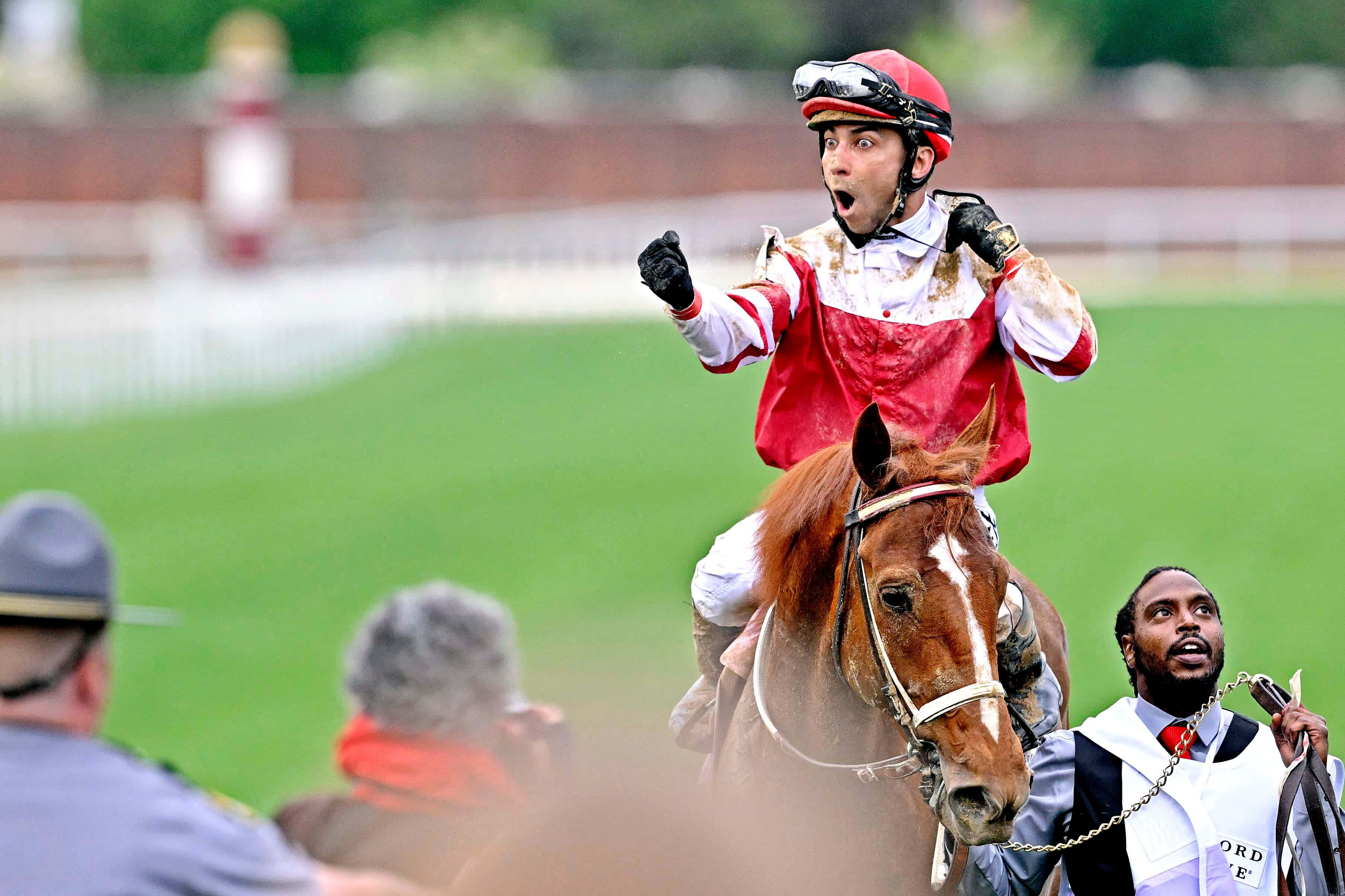 Kentucky Derby 桌布 壁紙 — 一位身穿紅色絲綢賽衣的騎師在賽馬場上騎著栗色馬匹瘋狂慶祝，觀眾在旁觀看。