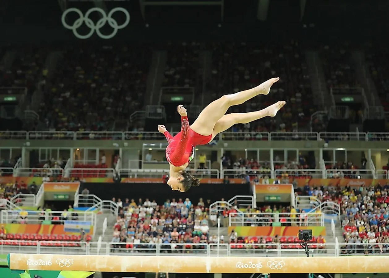 Olympics 桌布 壁紙 — 一位身穿紅色緊身衣的女子體操運動員在里約2016奧運會明亮的場館燈光下，在平衡木上完美地完成了一個後空翻。