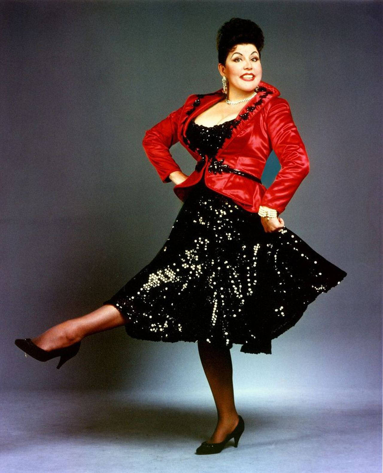 Singer 桌布 壁紙 — 一位身穿紅色緞面外套和閃亮黑色連衣裙的魅力女性，在攝影棚背景下擺出自信的姿勢。