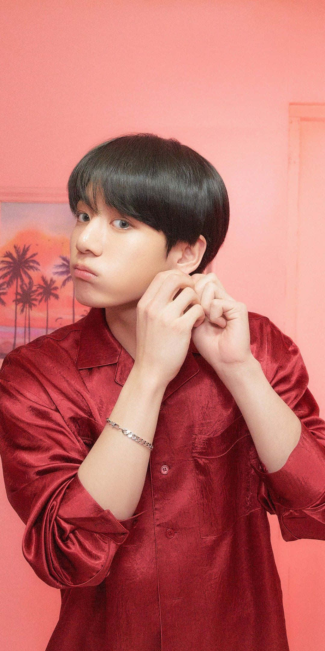 Jungkook 桌布 壁紙 — 一位身穿紅色緞面襯衫的年輕男子，將耳朵貼在粉紅色牆壁上調整，身後有一幅熱帶日落畫作。