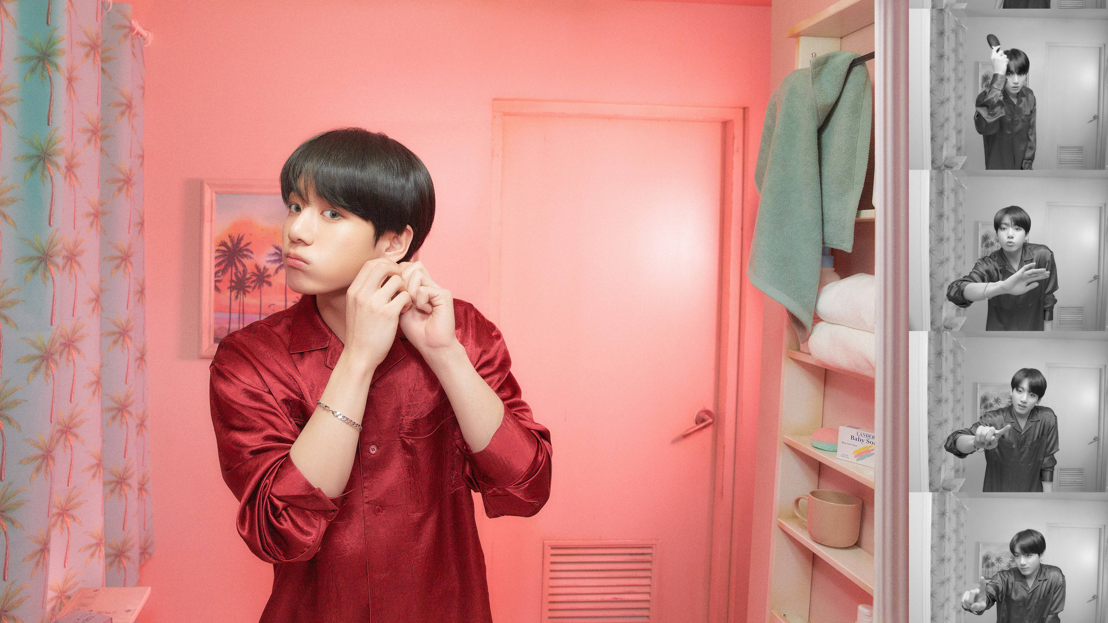 Jungkook 桌布 壁紙 — 一位身穿紅色緞面襯衫的年輕男子在一個粉紅色房間裡調整耳機，房間有棕櫚圖案的窗簾和一個裝滿毛巾的衣櫃。