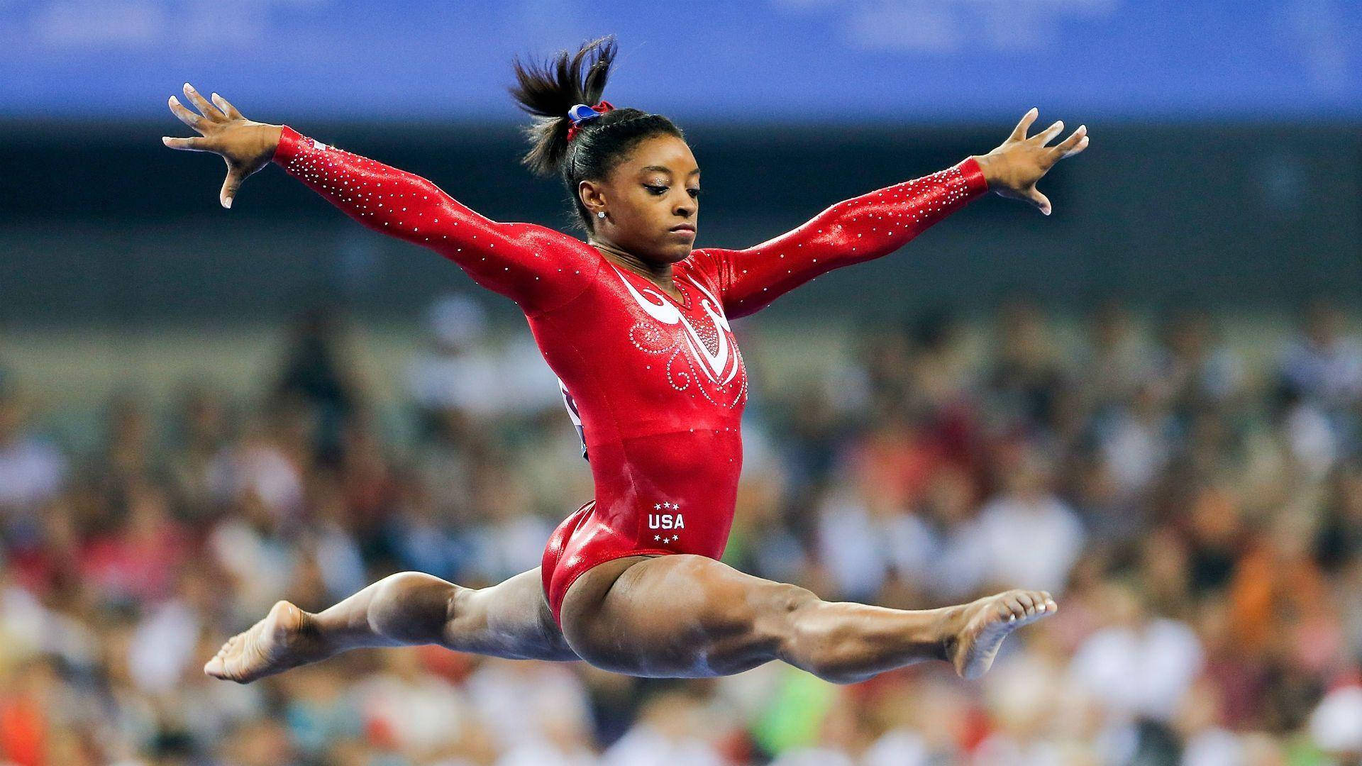 Simone Biles 桌布 壁紙 — 一位身穿紅色美國體操服的女體操運動員在比賽中於空中執行分腿跳躍，背景是模糊的人群。