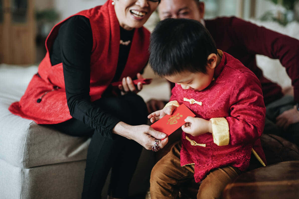 Red Envelope 桌布 壁紙 — 一位身穿紅色背心的快樂女士，在沙發上將一個紅包遞給一個穿著傳統紅色服裝的孩子。