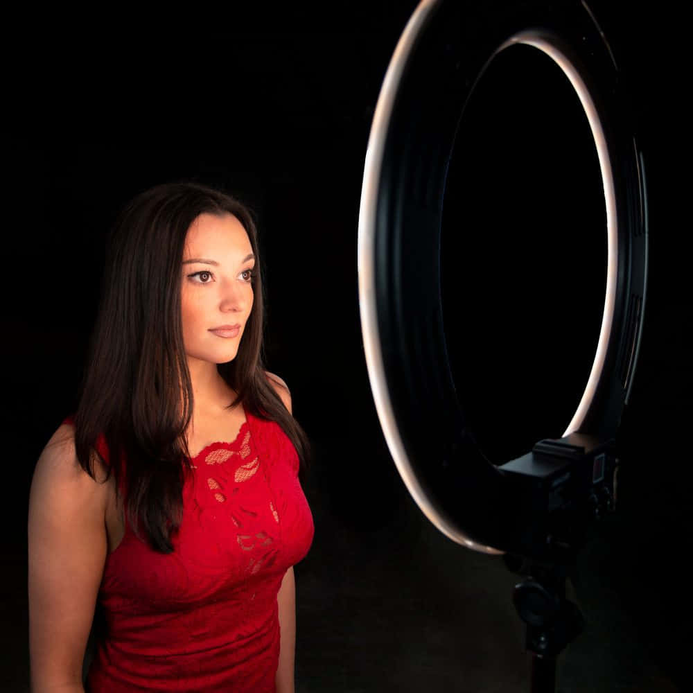 Ring Light 桌布 壁紙 — 一位身穿紅色蕾絲上衣的女性站在發光的環形燈旁，背景為黑色。