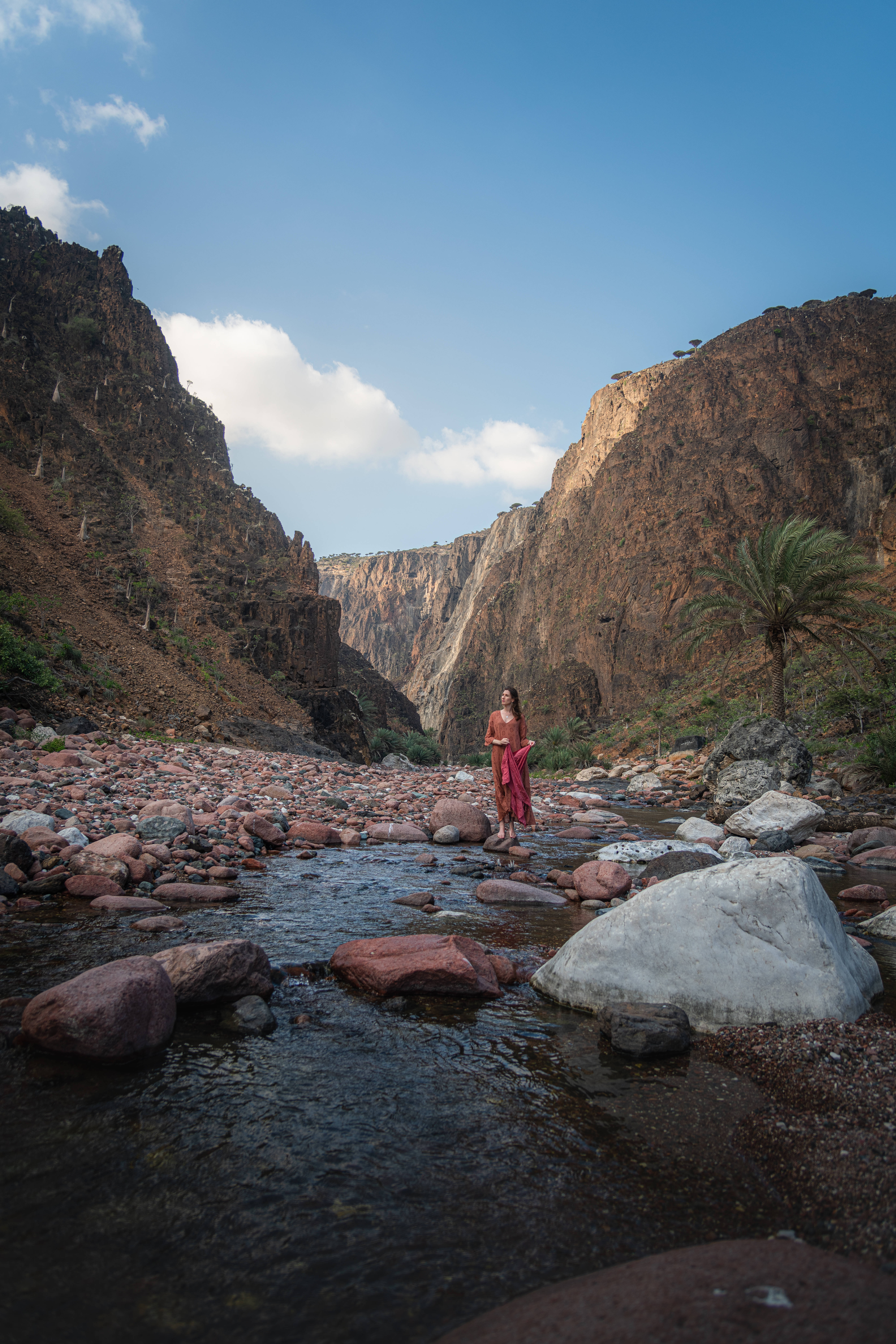 Yemen 桌布 壁紙 — 一位身穿紅色衣服的女性赤腳站在淺峽谷溪流中的岩石上，頭頂是湛藍的天空，點綴著幾朵白雲。