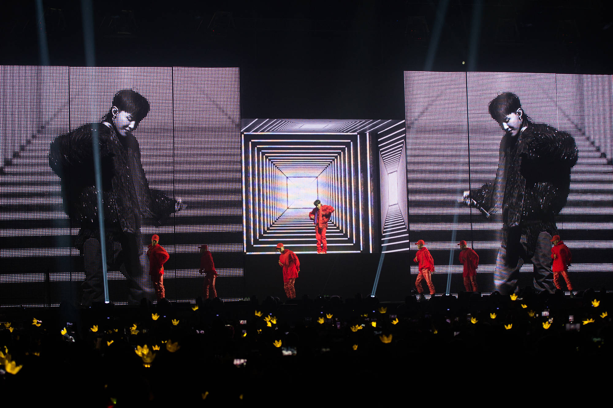 G-dragon 桌布 壁紙 — 一位身穿紅色表演者站在舞台中央，處於發光的數位隧道內，兩旁的大螢幕顯示他的剪影，紅色舞者在他周圍移動，聚光燈下，觀眾在下方舉著黃色燈光。