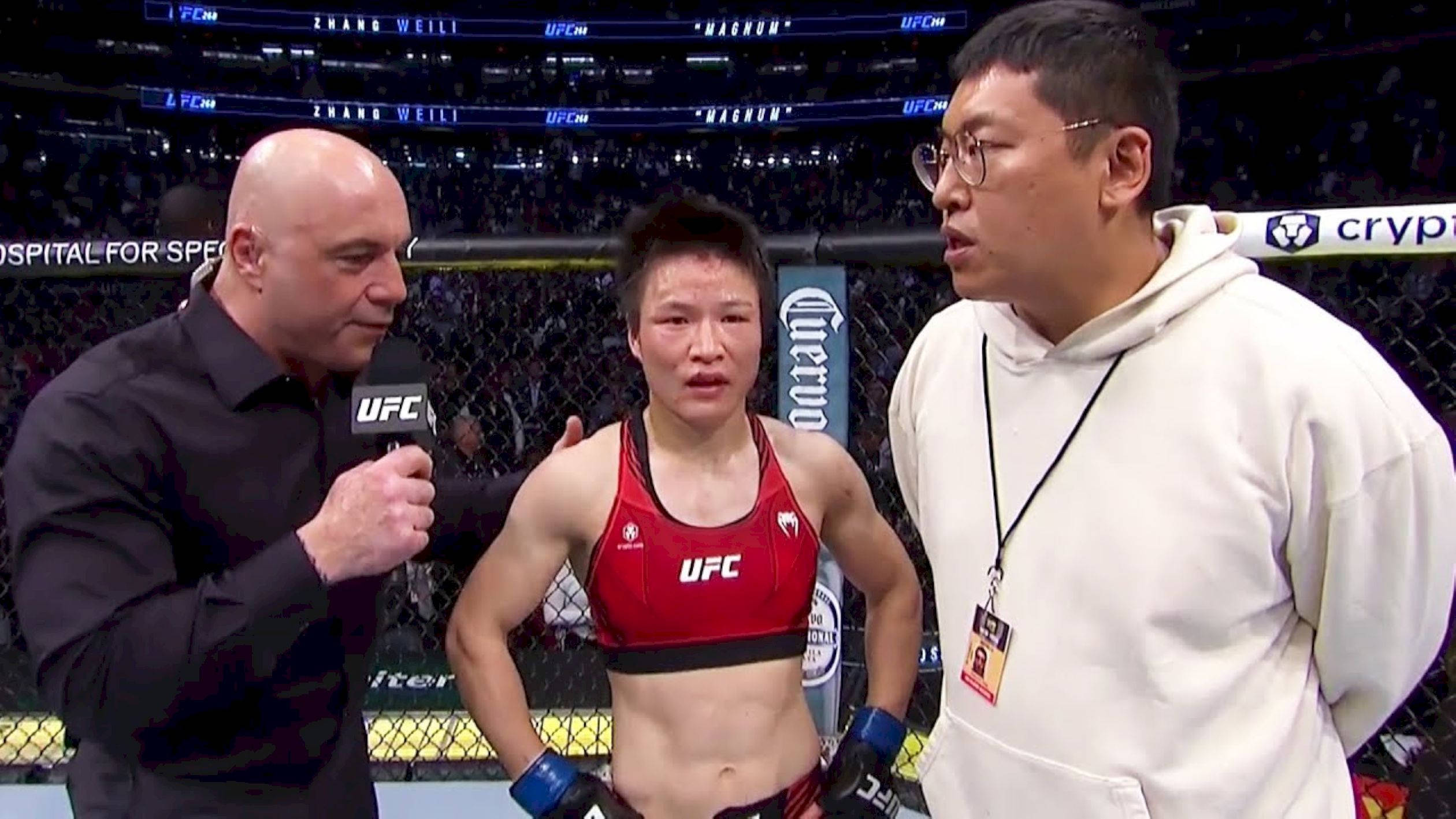 Athletic 桌布 壁紙 — 一位身穿紅色裝備的女性UFC拳手站在光頭採訪者和一位身穿白色連帽衫的男子之間，位於八角籠內。