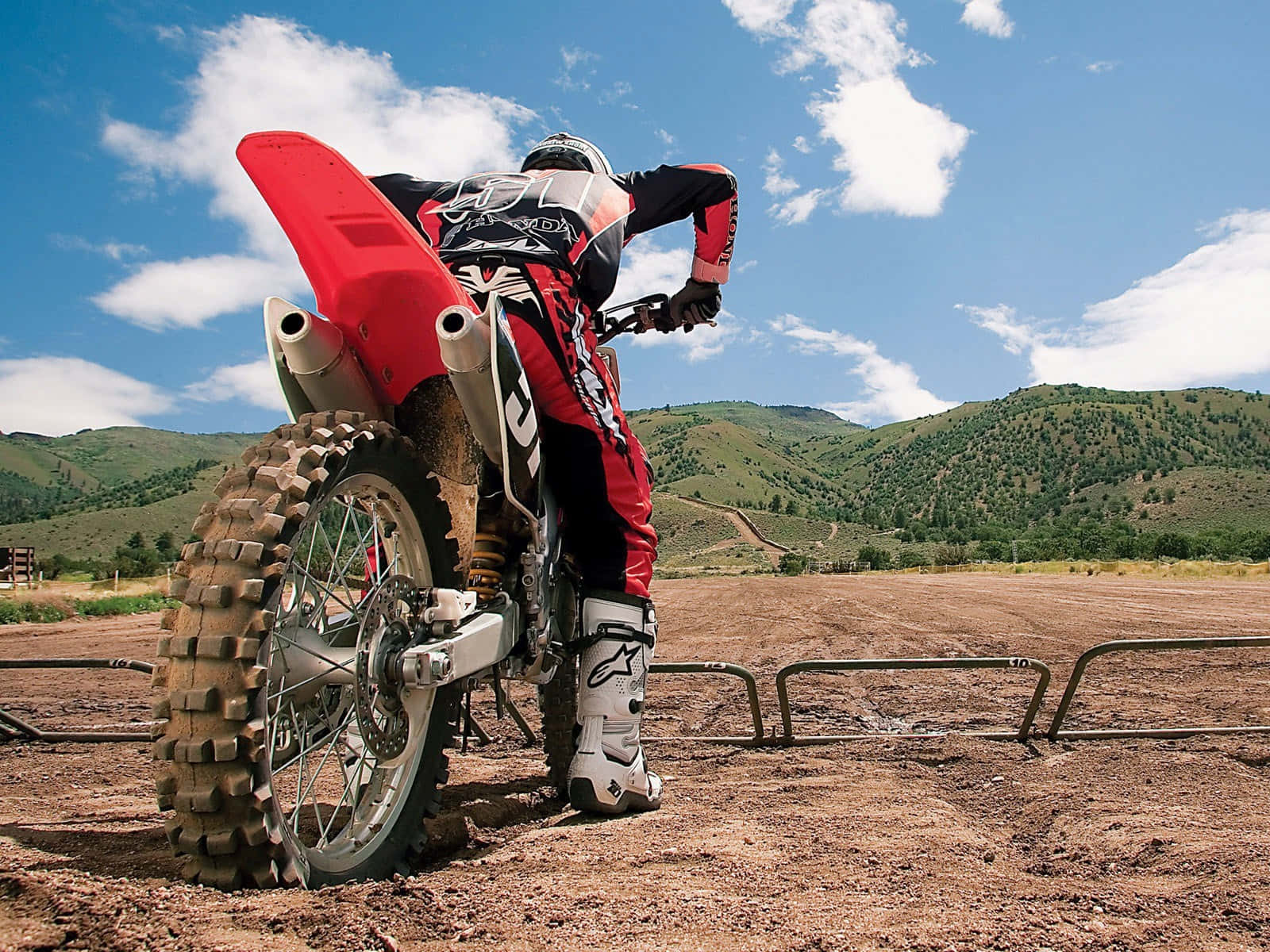 Motocross 桌布 壁紙 — 一位身穿紅色裝備的越野摩托車手在 dirt bike 上傾斜入彎，背景是起伏的綠色山丘和藍天。