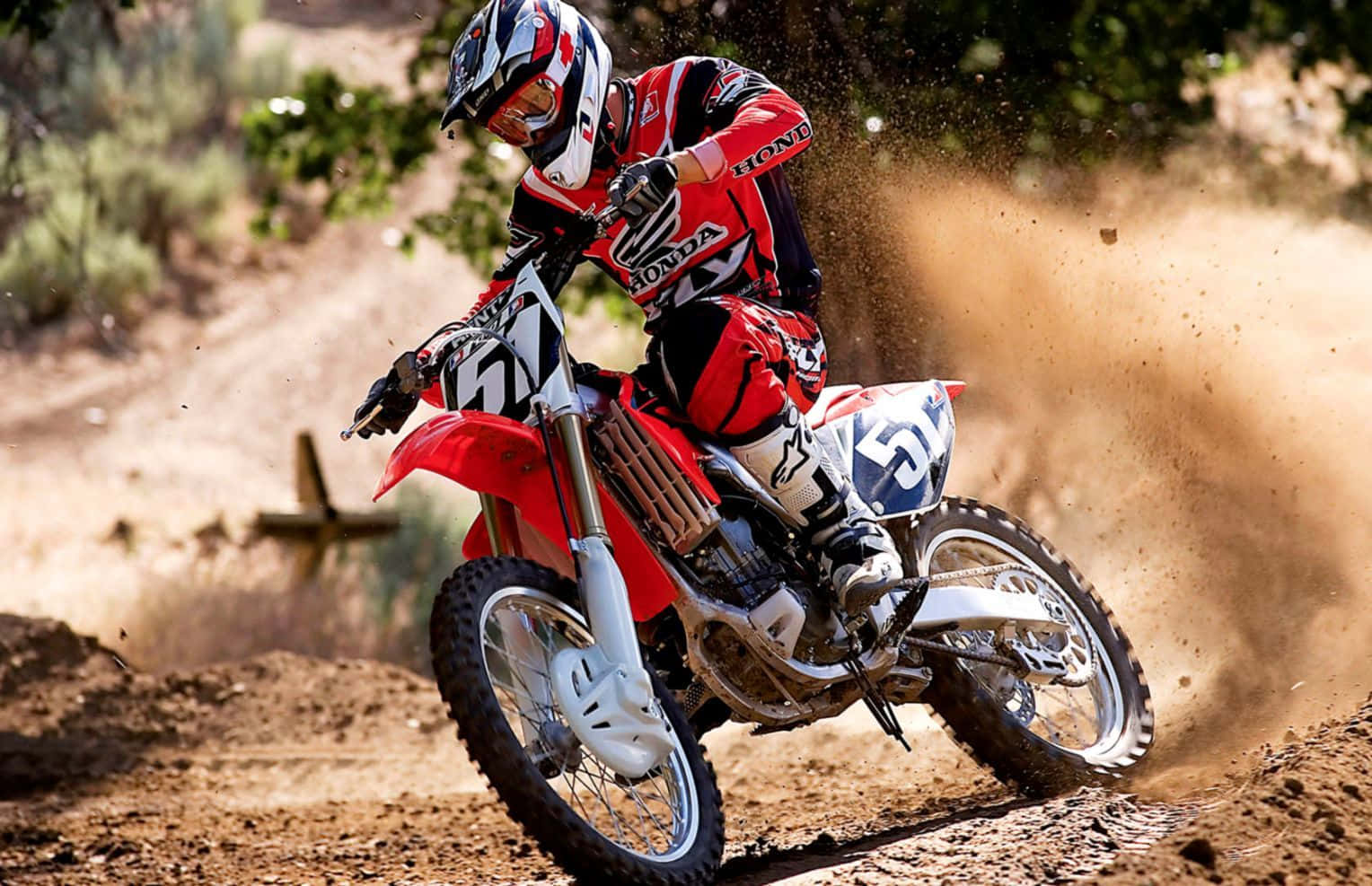 Honda Dirt Bike 桌布 壁紙 — 一位身穿紅色裝備的越野機車手在紅色本田越野機車上傾斜入彎，在陽光明媚的泥地賽道上揚起灰塵。