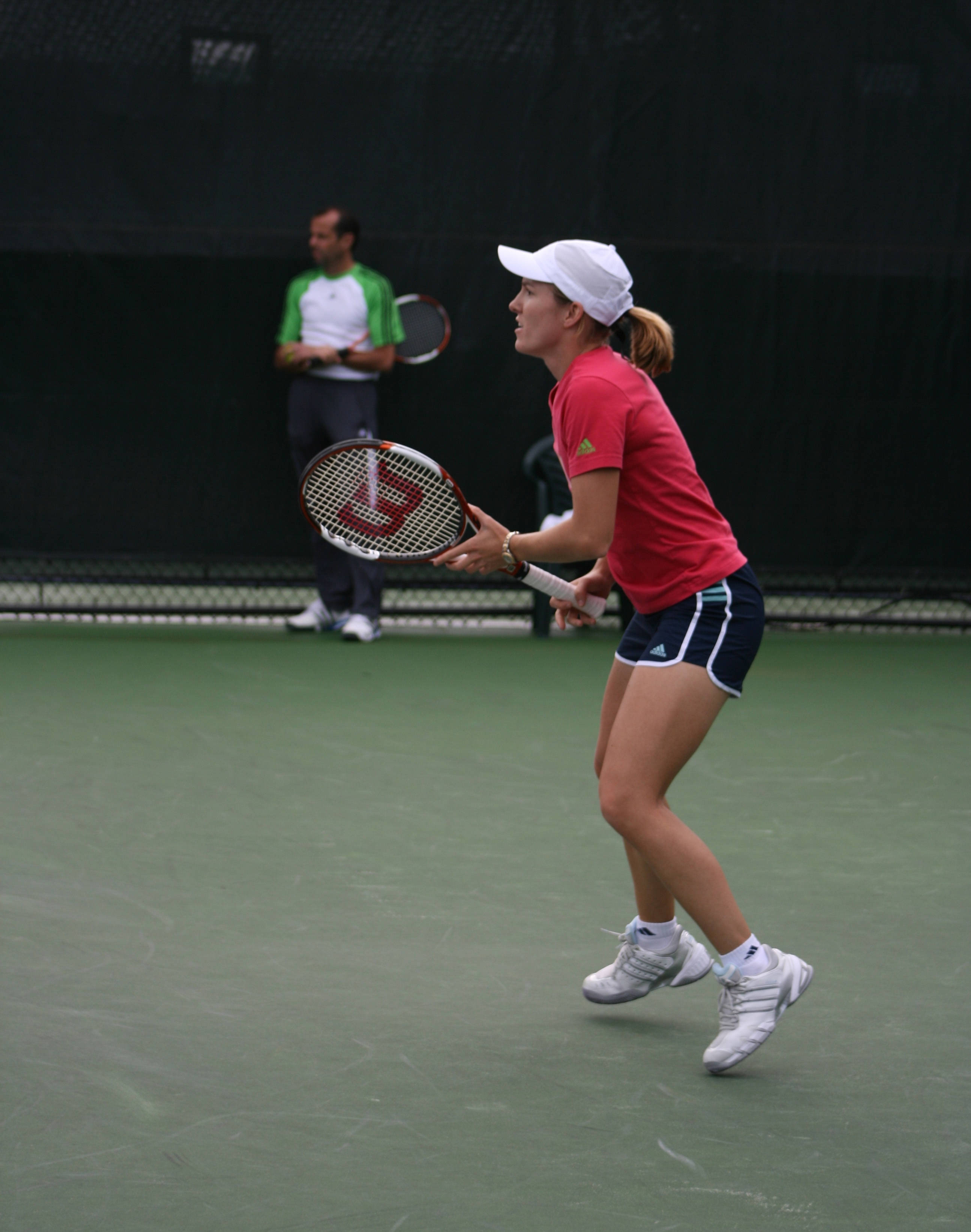 Tennis 桌布 壁紙 — 一位身穿紅色襯衫和白色帽子的女網球運動員在綠色球場上準備正手擊球，教練在背景中觀看。