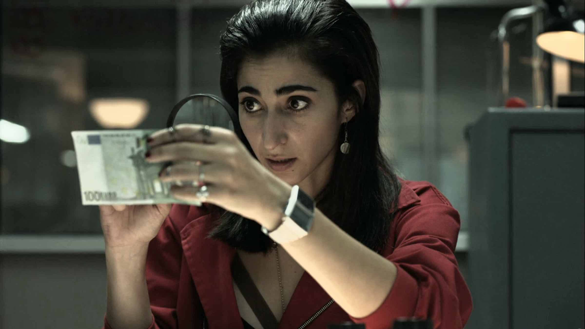 nairobi money heist 桌布 壁紙 — 一位身穿紅色襯衫的女性在燈光昏暗的辦公室裡透過放大鏡檢查一張100歐元紙幣。