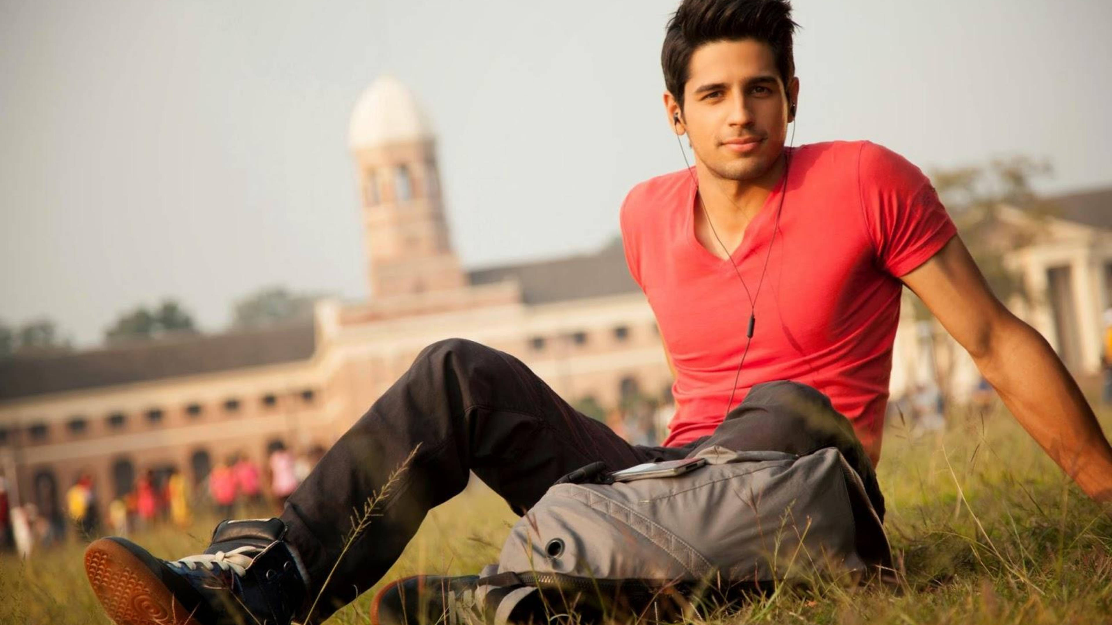 Sidharth Malhotra 桌布 壁紙 — 一位身穿紅色襯衫的年輕男子背著背包，戴著耳機，以帶有圓頂的歷史建築為背景，放鬆地坐在草地上。