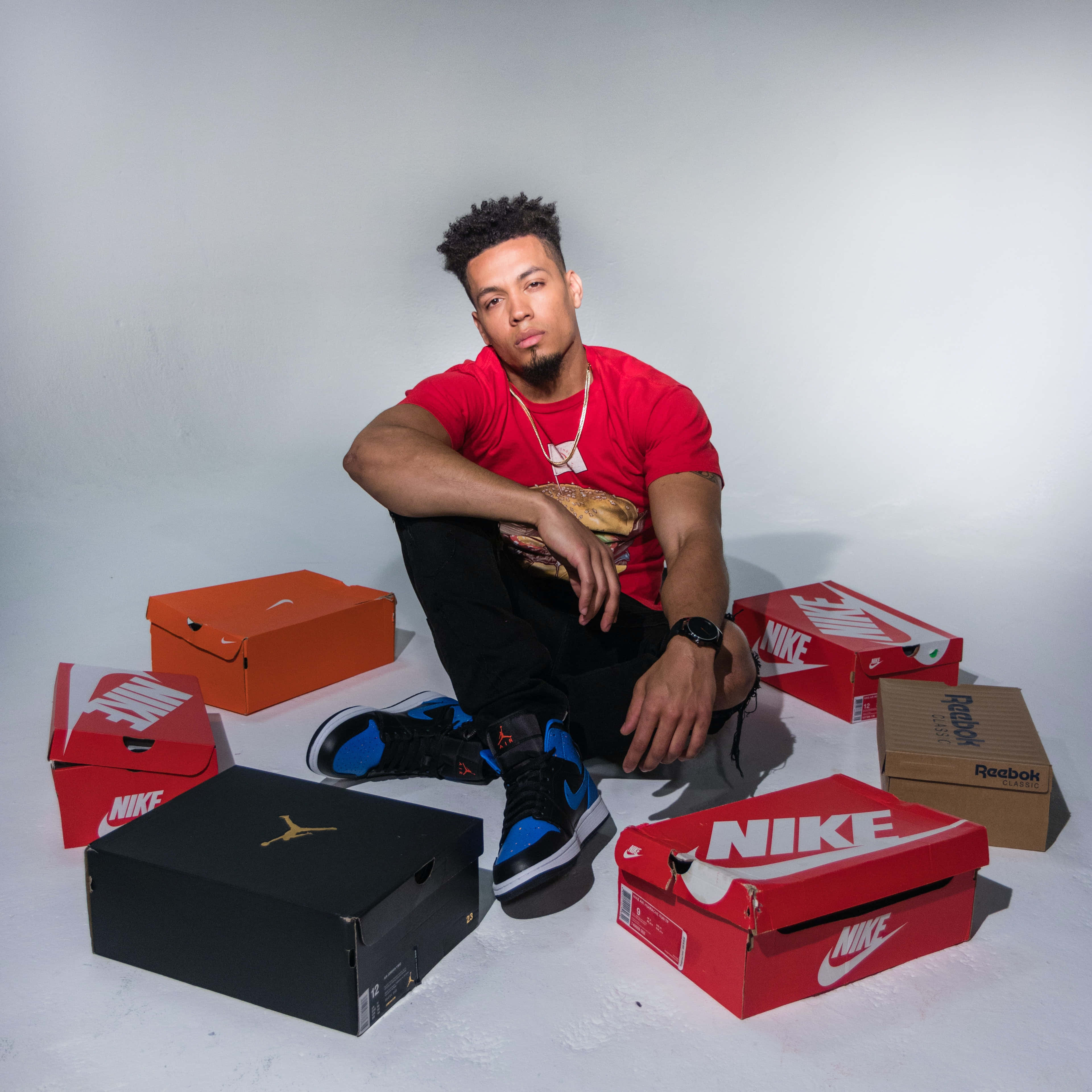 Shoe Box 桌布 壁紙 — 一位身穿紅色襯衫的年輕男子自信地坐在白色地板上，周圍是色彩繽紛的運動鞋盒，背景是素牆。