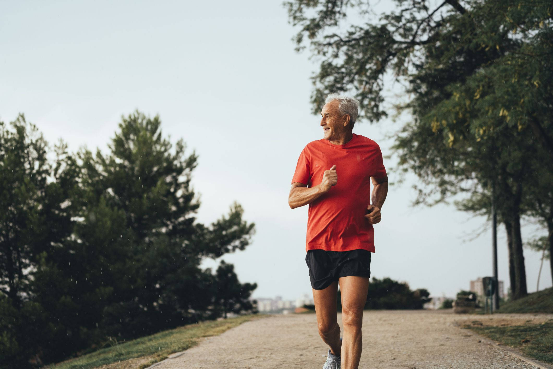 Running 桌布 壁紙 — 一位身穿紅色襯衫的年長男性在多雲的天空下，沿著綠樹成蔭的小徑充滿活力地奔跑。
