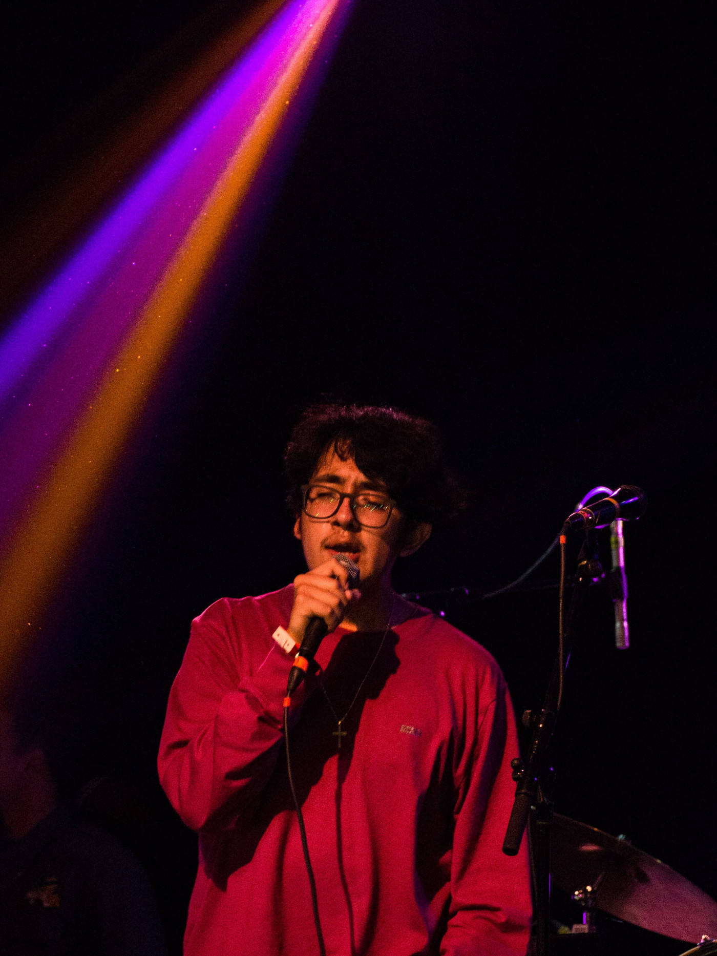Singing 桌布 壁紙 — 一位身穿紅色襯衫的音樂家在戲劇性的紫色和橙色舞台燈光下對著麥克風唱歌，背景為深色。